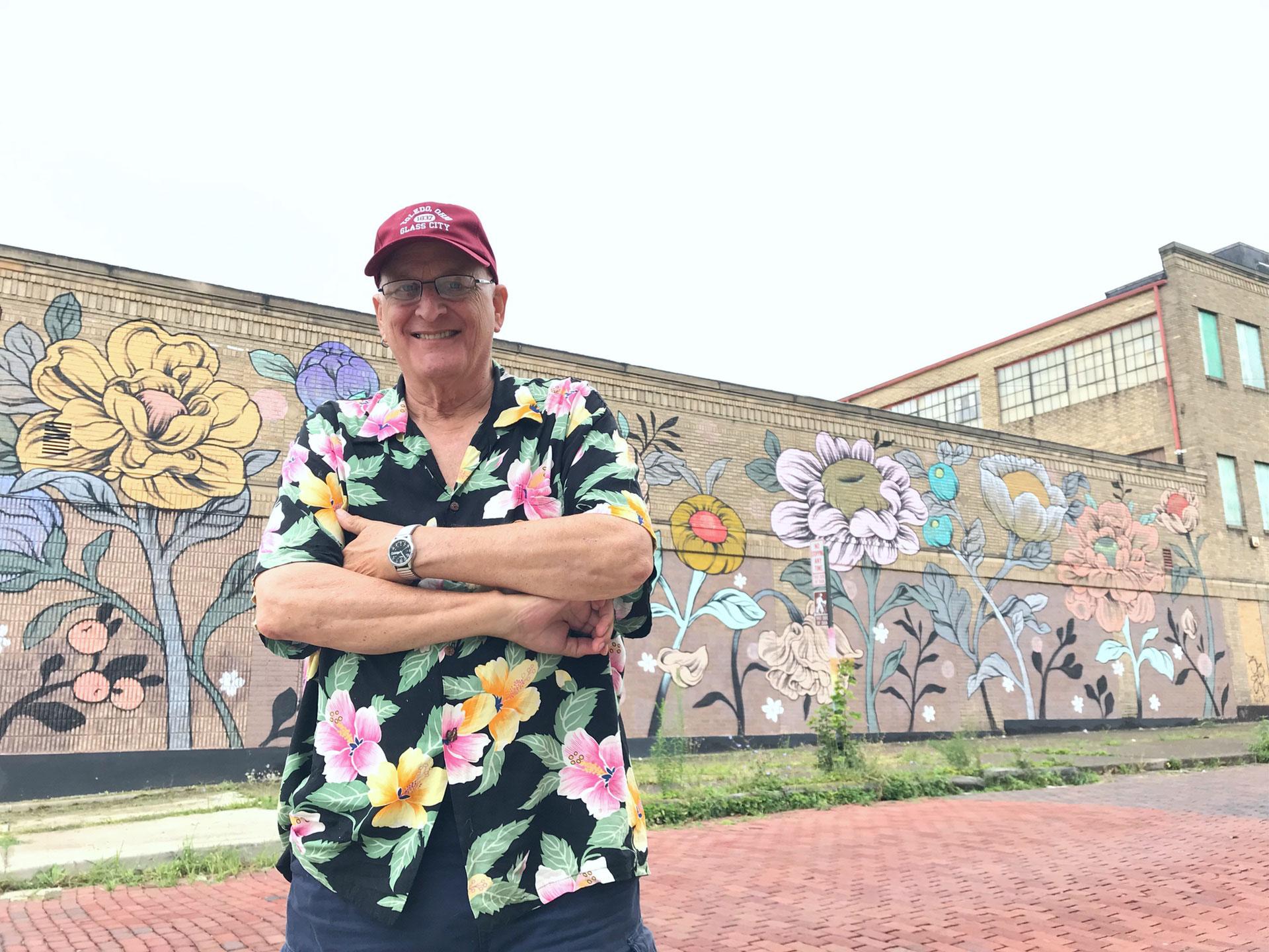 A man is shown wearing a flower-print shirt and red hat while standing in front of a wall with flowers painted on it.