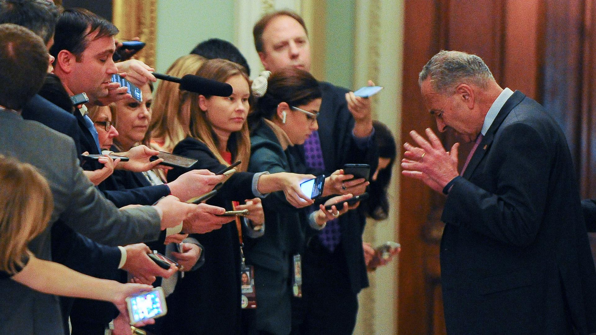 US Senate Minority Leader Chuck Schumer is shown with his hands raised and head down with a row of reporters standing across from him.