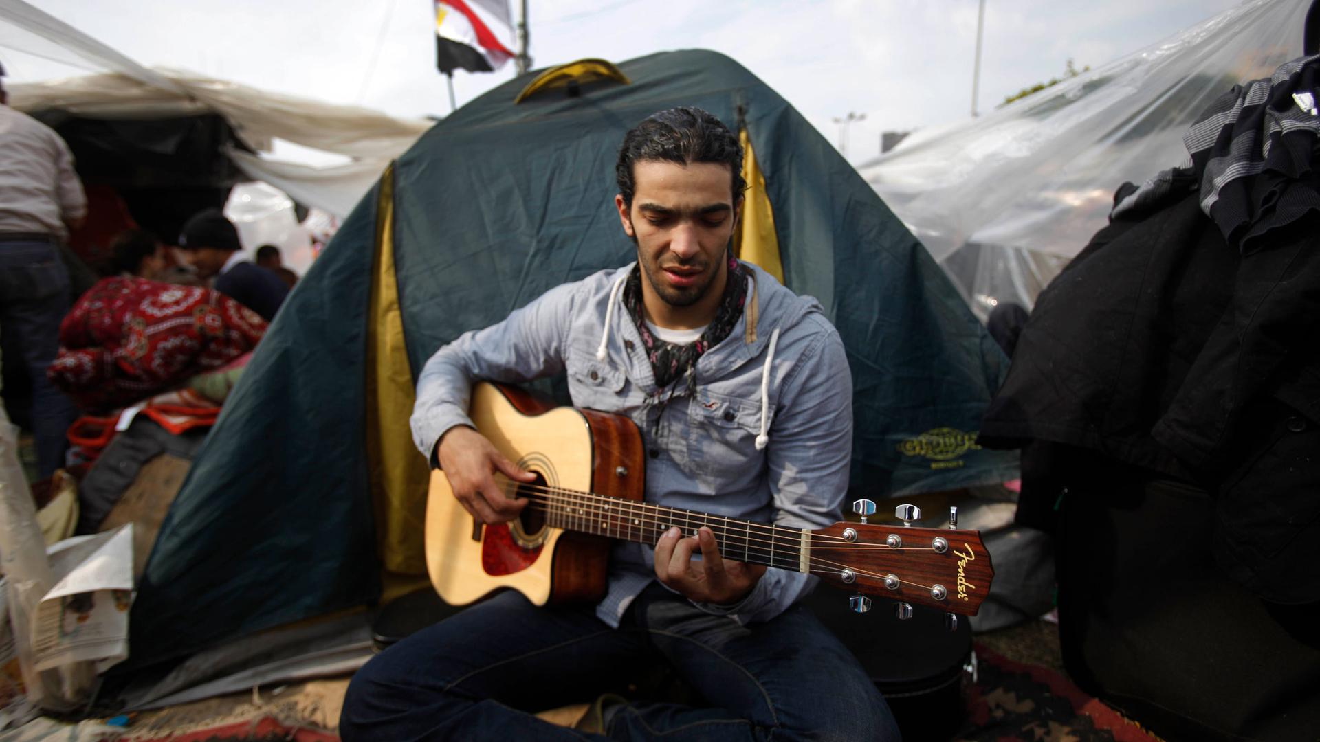 A man is sitting cross-legged in front of a tent and holding a guitar.