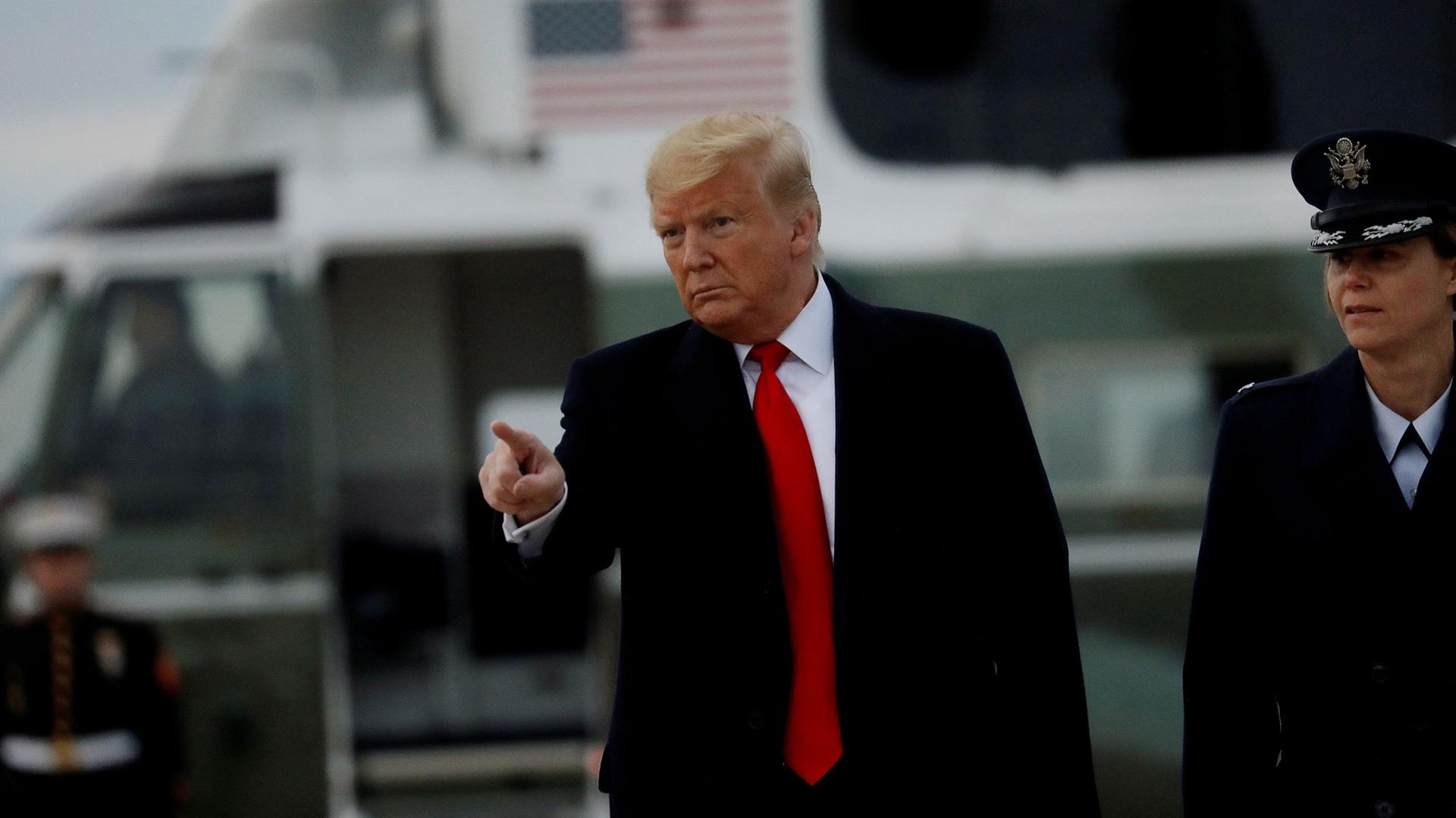 US President Donald Trump is shown pointing with his right hand and wearing a dark jacket and red tie.