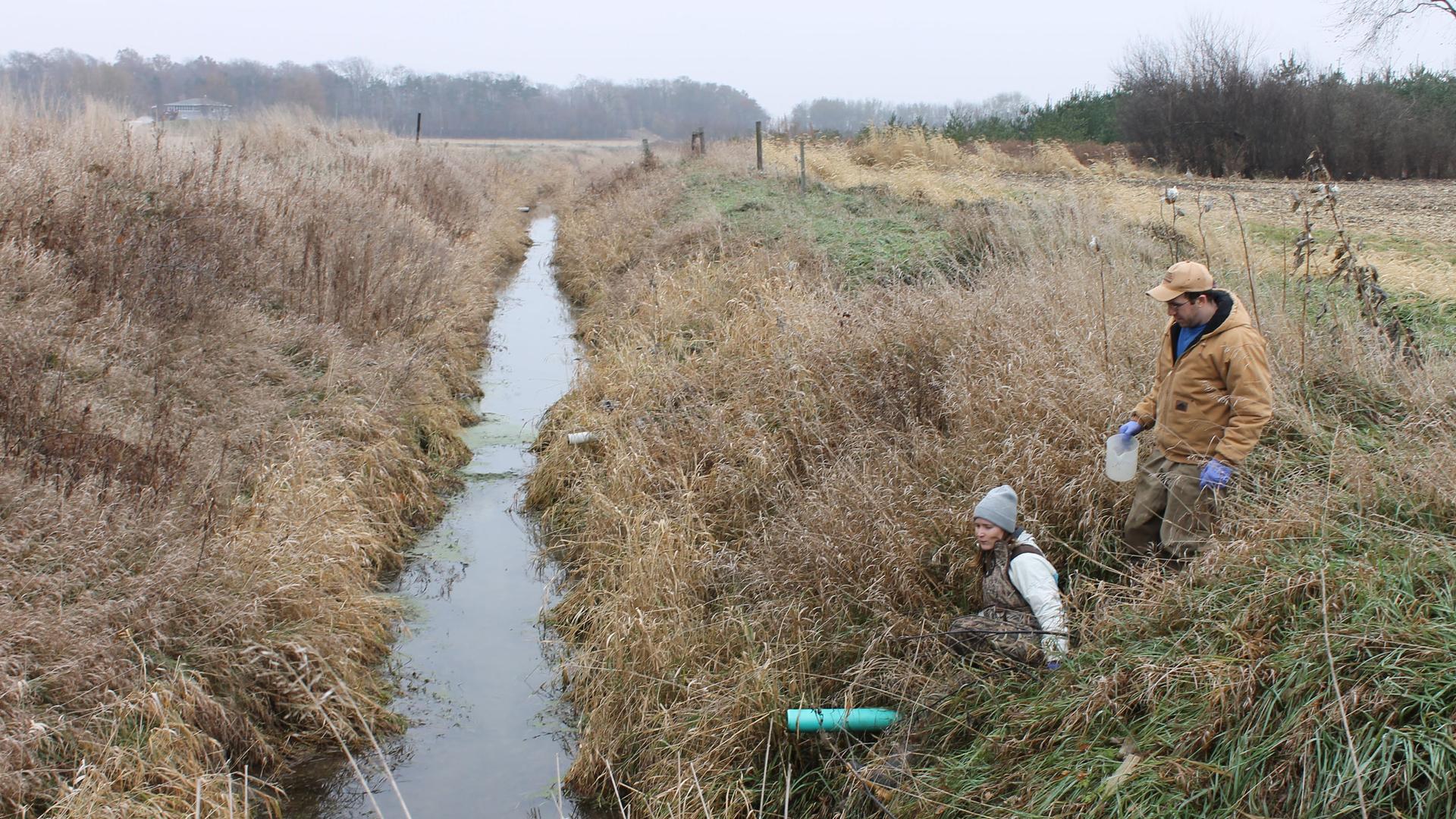 Two people are shown next to a water-filled ditch with a grassy field on either side.