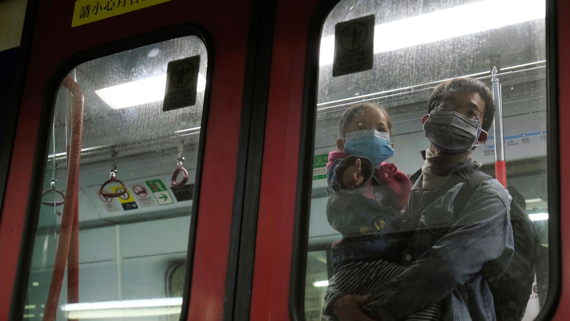 A man is shown holding a child, both wearing face masks, and shown through the window of a train.