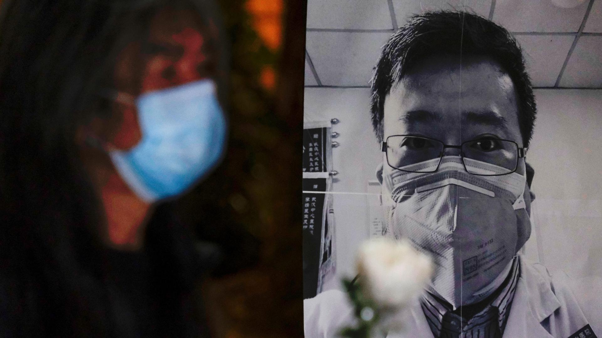 A person wearing a mask is shown in soft focu near a black and white photograph of Dr. Li Wenliang.