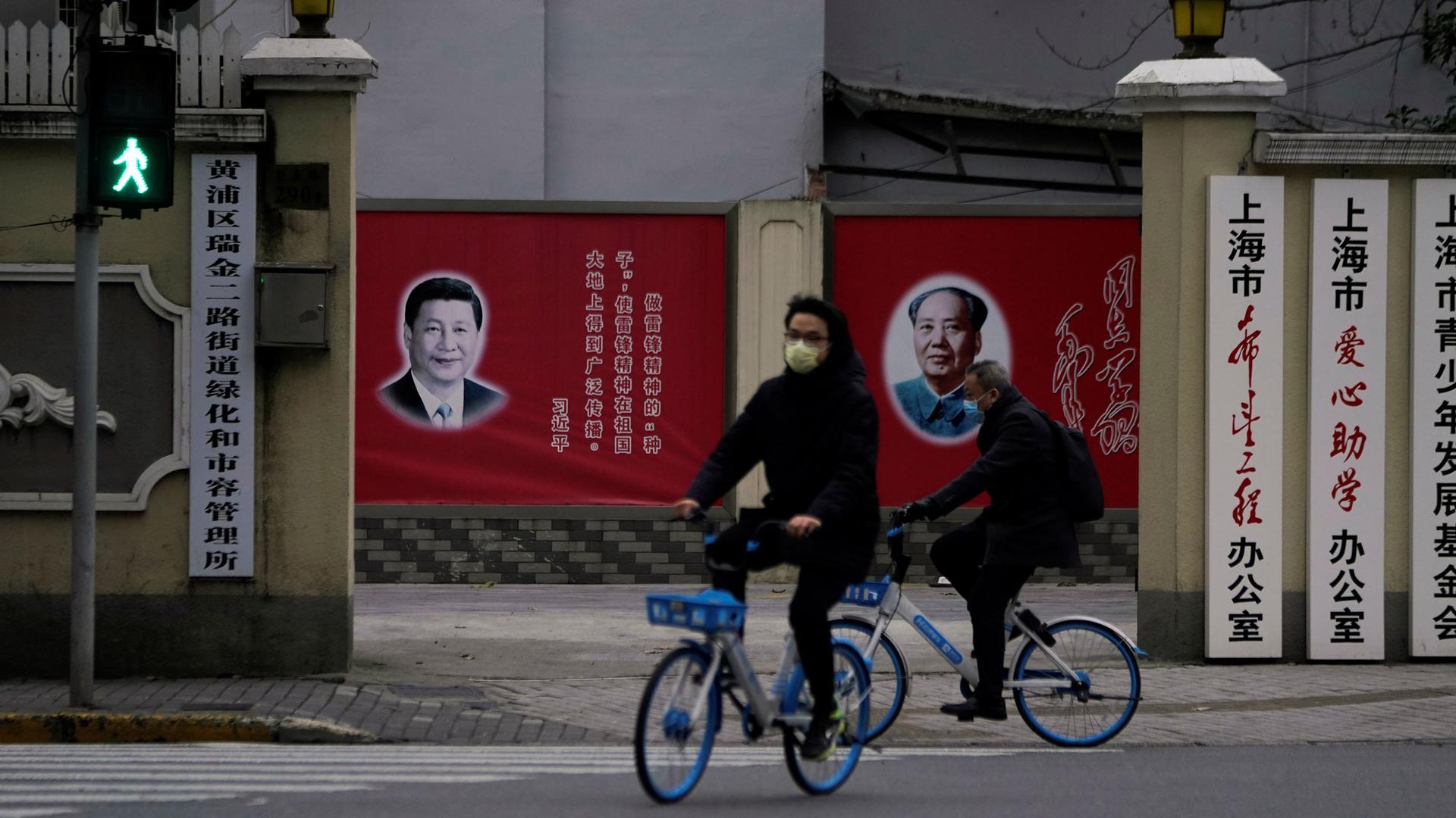 Two people riding bicycles are shown wearing face masks and passing by portraits of Chinese President Xi Jinping and late Chinese chairman Mao Zedong.
