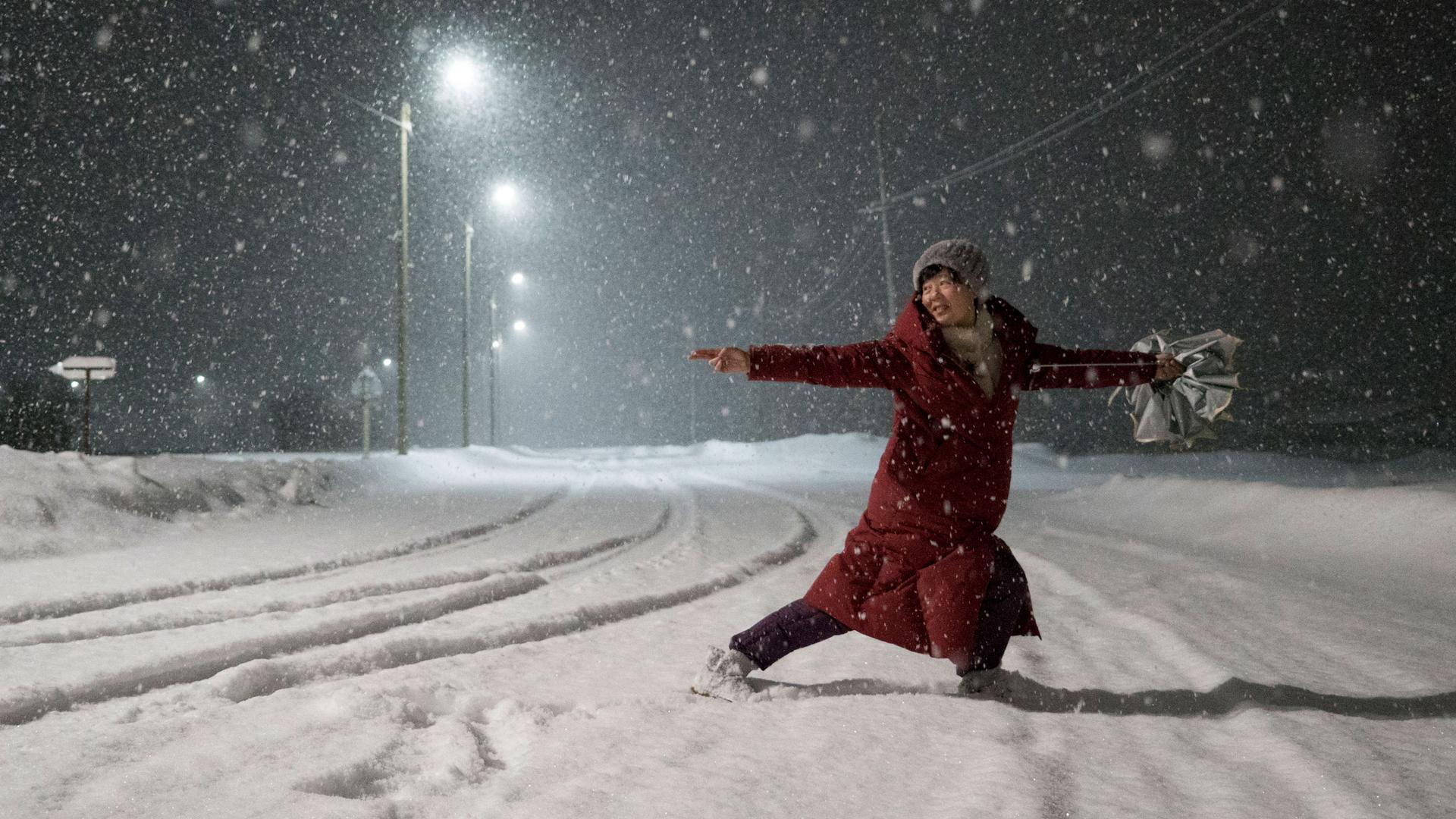 A woman in a red jacket is shown dancing in the street with snow falling all around her.