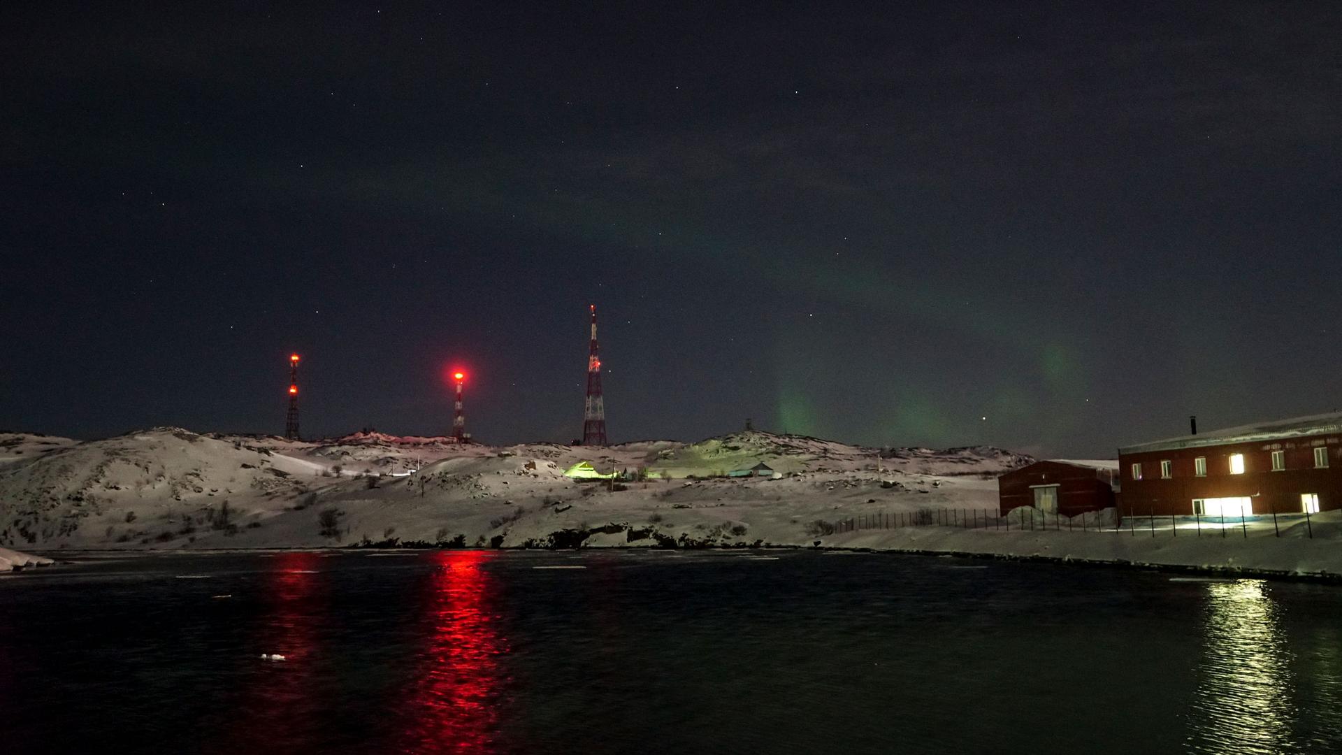 A green swirl of the northern lights is shown against a dark sky with antennas with red lights in the nearground.