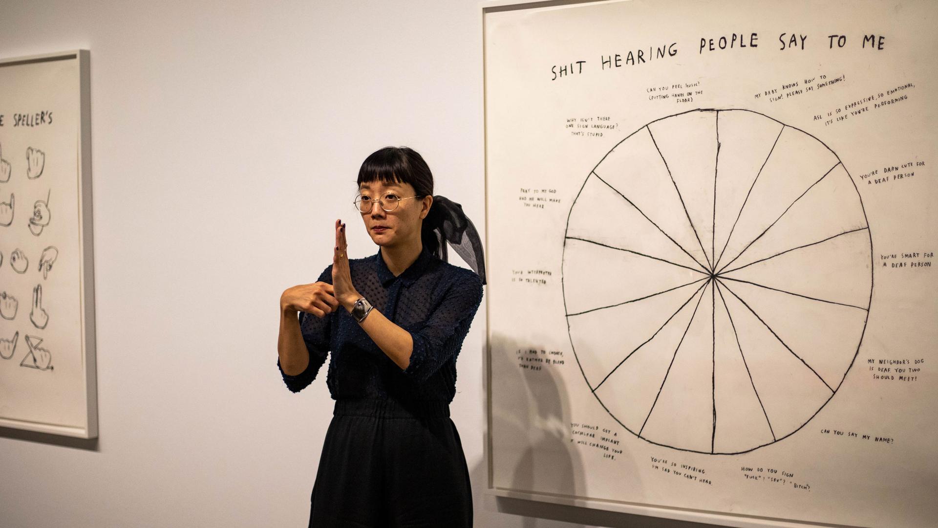 Christine Sun Kim is shown signing and standing in front of her art featuring a pie chart titled "Shit Hearing People Say To Me."