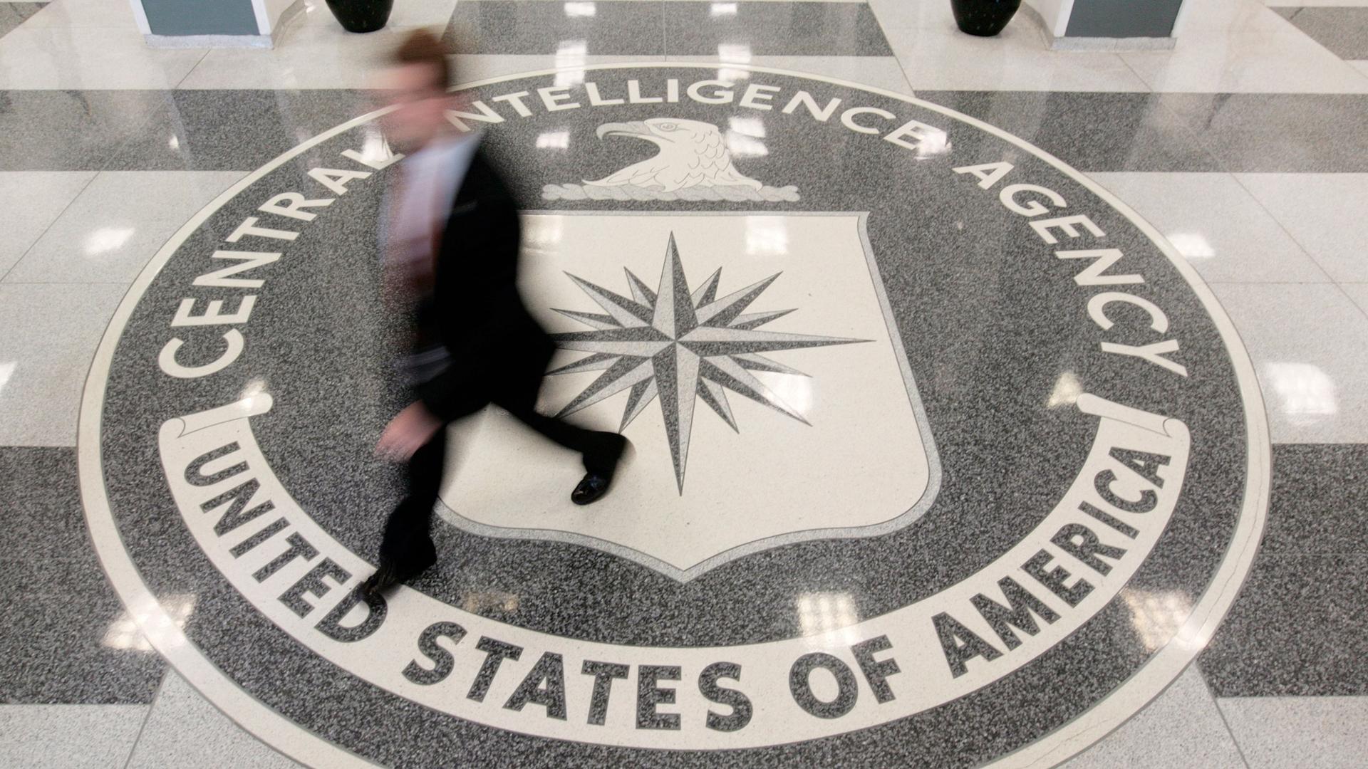 A seal of the CIA is shown from above with a person walking over it in blurred focus.