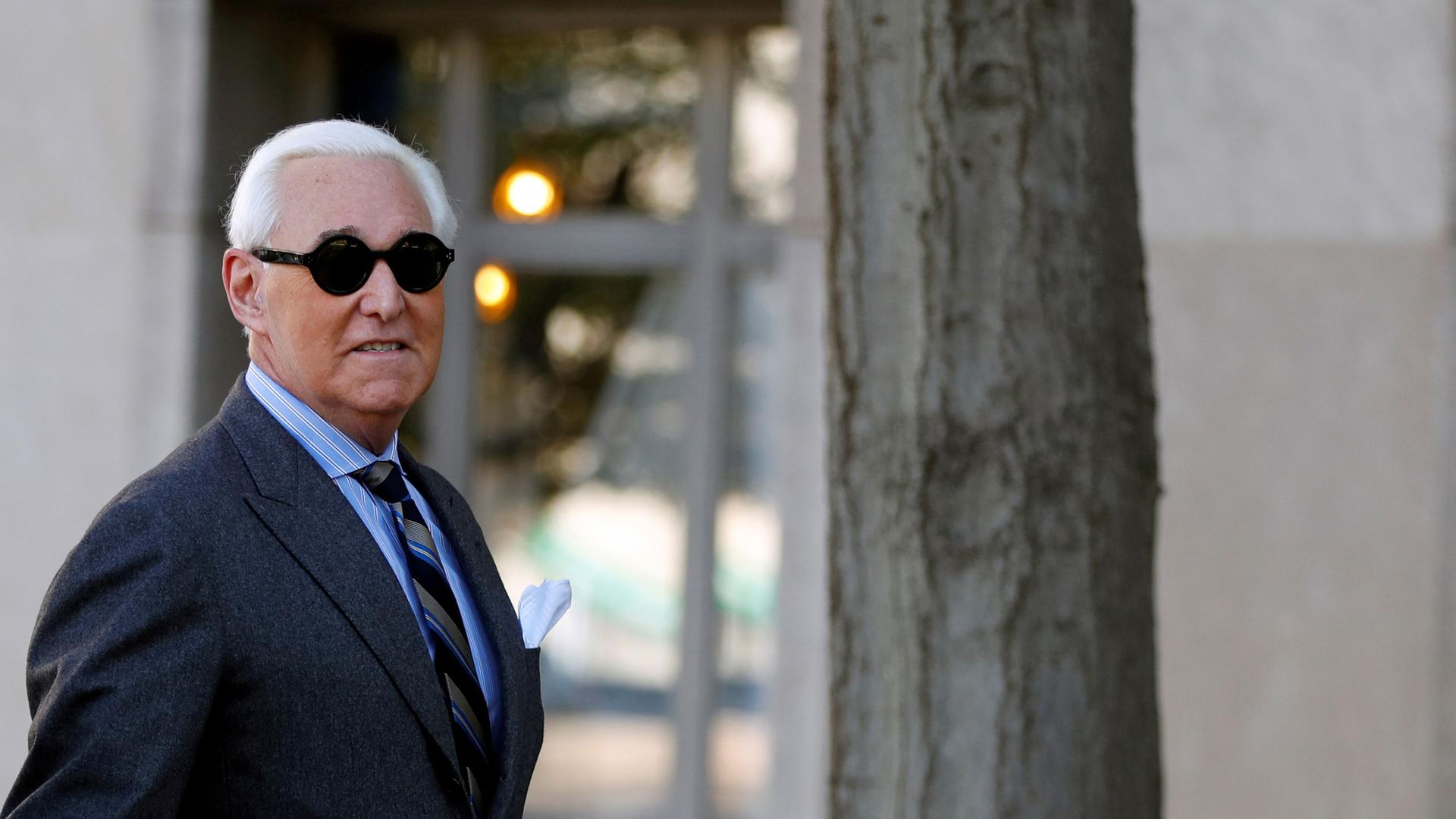 Roger Stone is shown wearing a grey suit and striped tie and dark circular sun glasses.