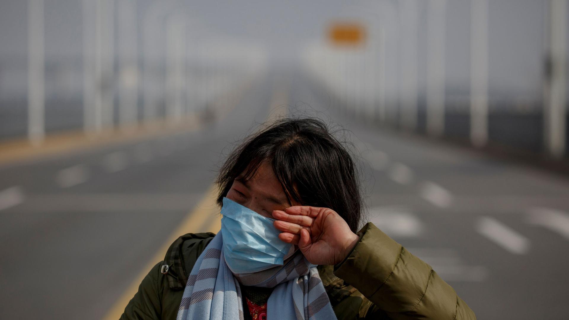 A woman is shown wearing a face mask and stripped scarf while standing on the edge of an empty bridge.