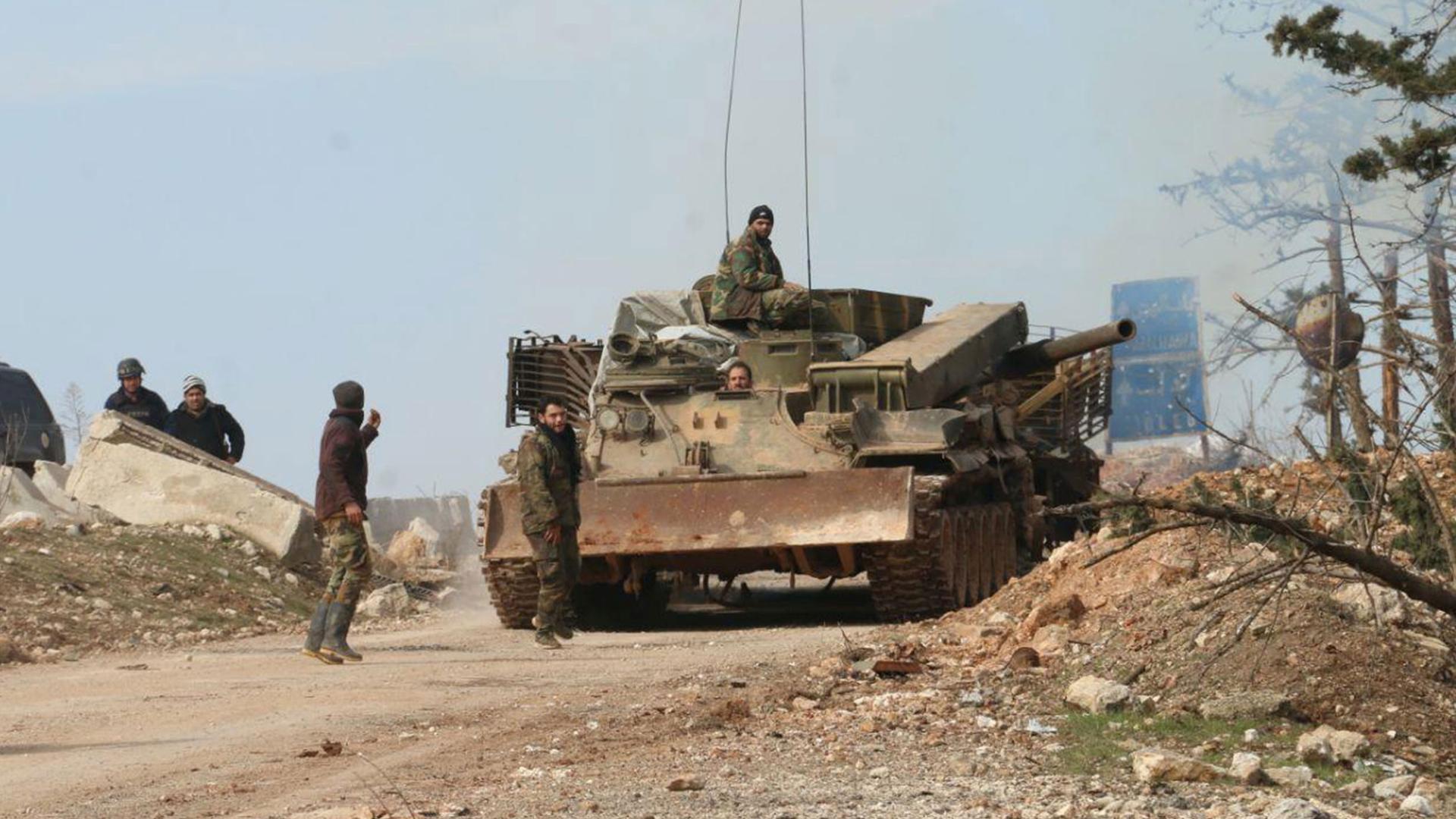Several Syria soldiers are shown walking alongside and riding on a tank in a dirt road.