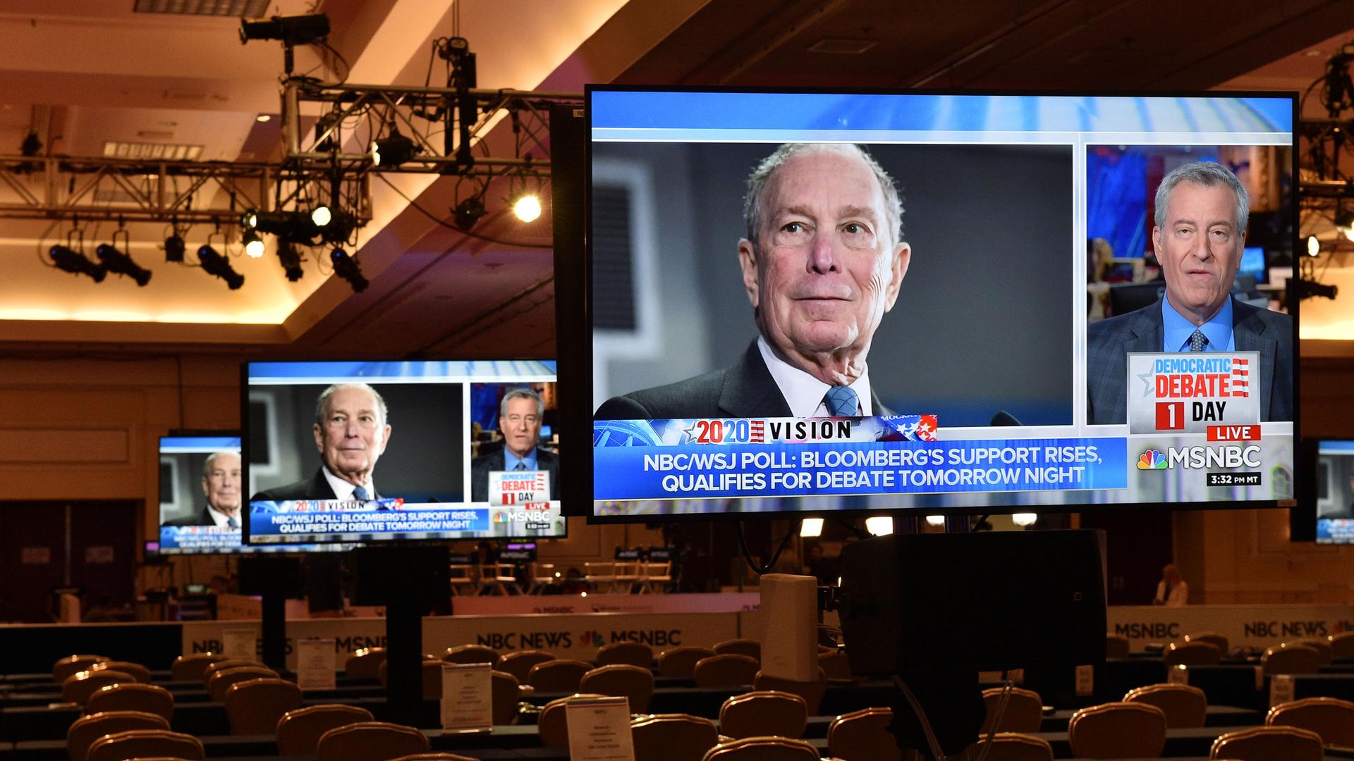 An image of the face of Michael Bloomberg is shown on a large video monitor in an auditorium with empty seats.