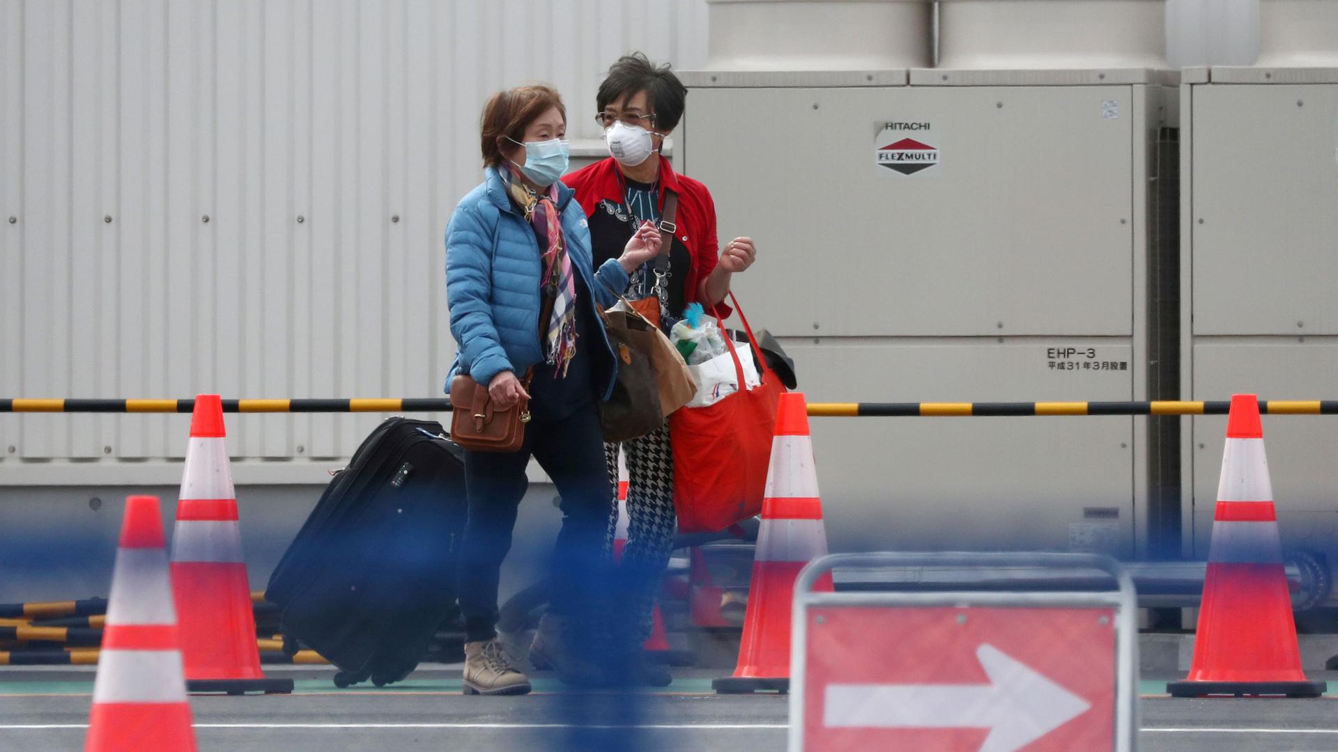 Two women are shown exiting a cruise ship wearing face masks and pulling suitcases.