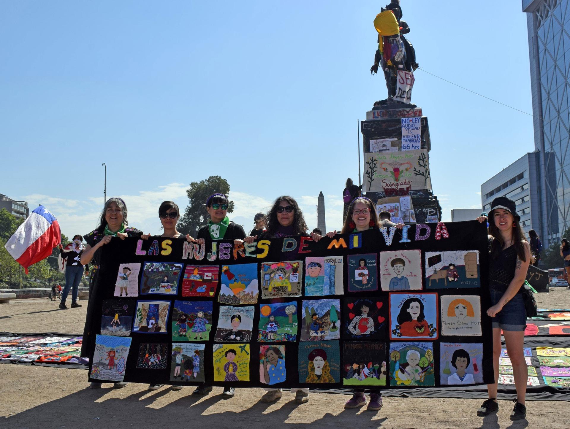 Several women are shown holding a tapestry with rows of embroidered blocks depicting slogans calling for legal abortion and an end to sexual assault.