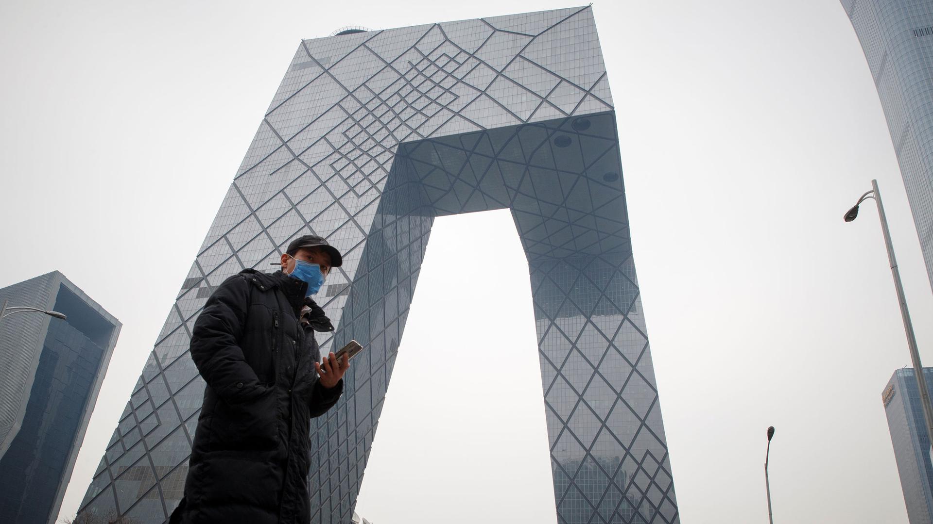A man is shown standing in front of the CCTV headquarters building with a mobile phone and wearing a face mask.