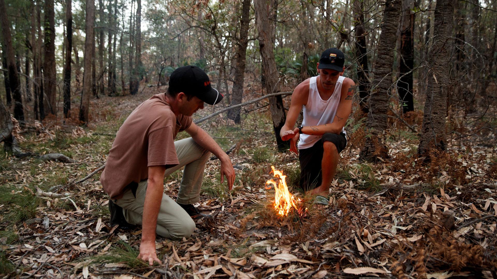 Two men bend over a small fire in the forest
