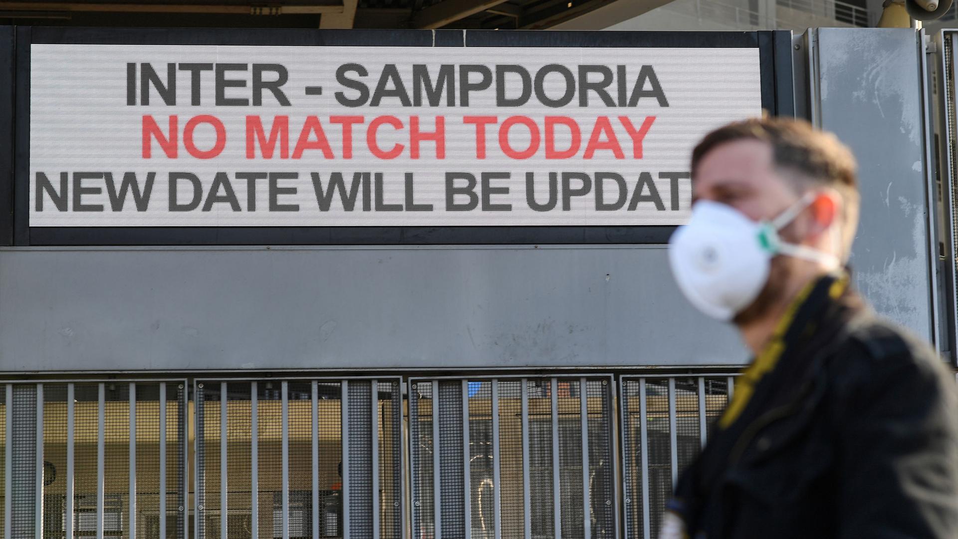 A man is shown in soft focus wearing a face mask and standing outside the San Siro stadium with a sign that reads, 