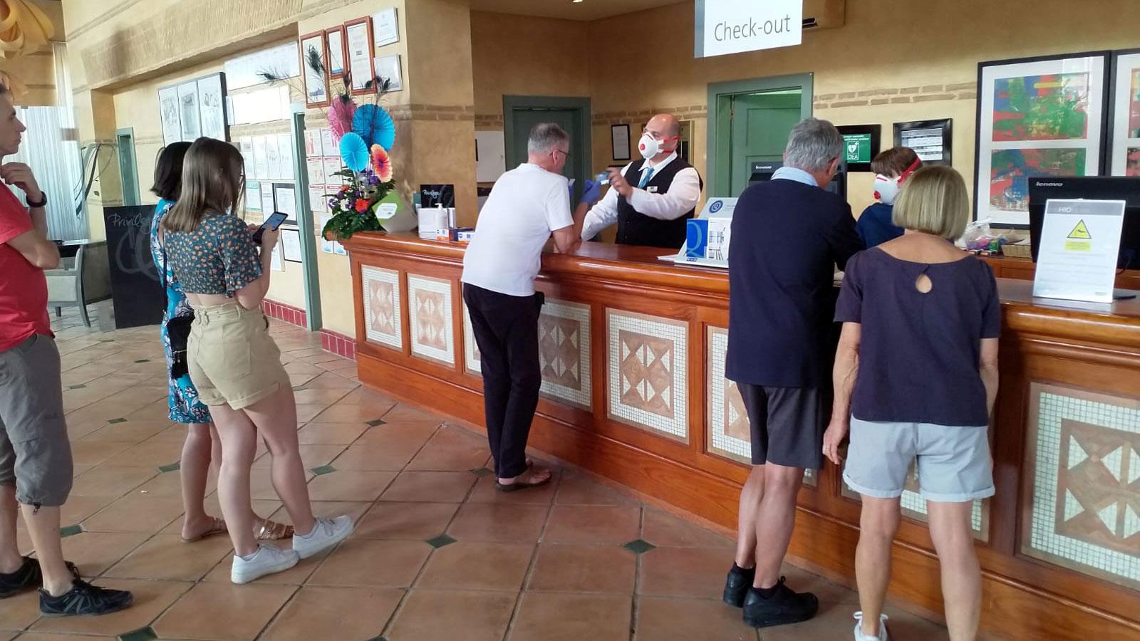 Several people are shown standing at the counter of a hotel resort with the words, "Check out" on a sign behind the counter.