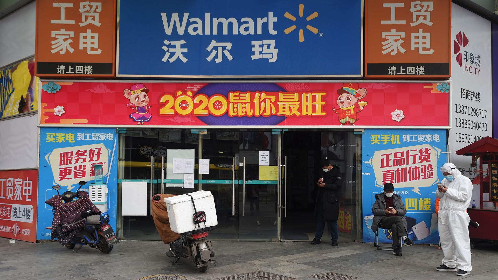 A colorful entrance to a Walmart in Wuhan, China, is shown with several advertisements and two men nearby wearing face masks.