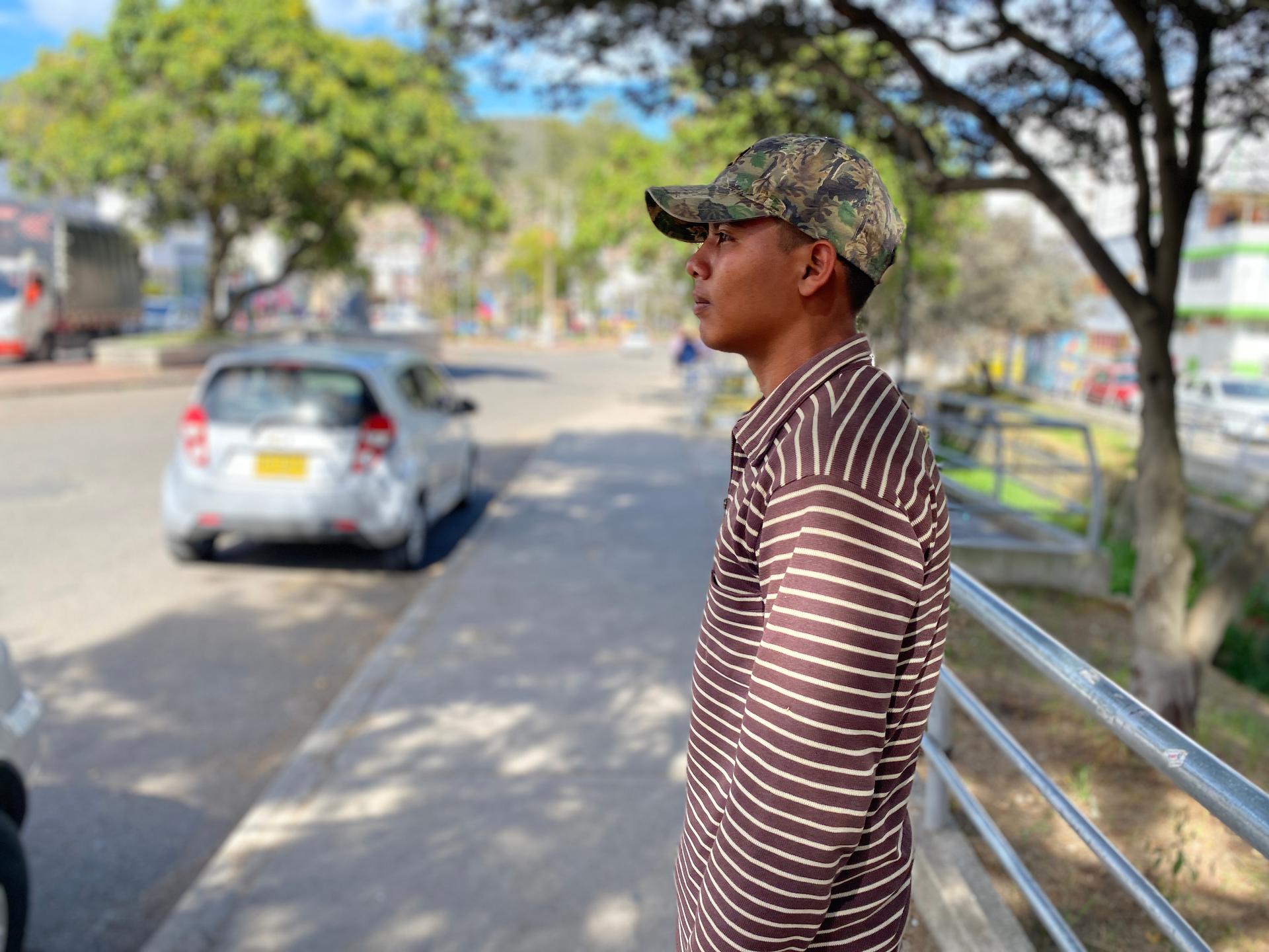 A young man wears a camouflage hat with brown and cream striped shirt and stands outside near a parked car.