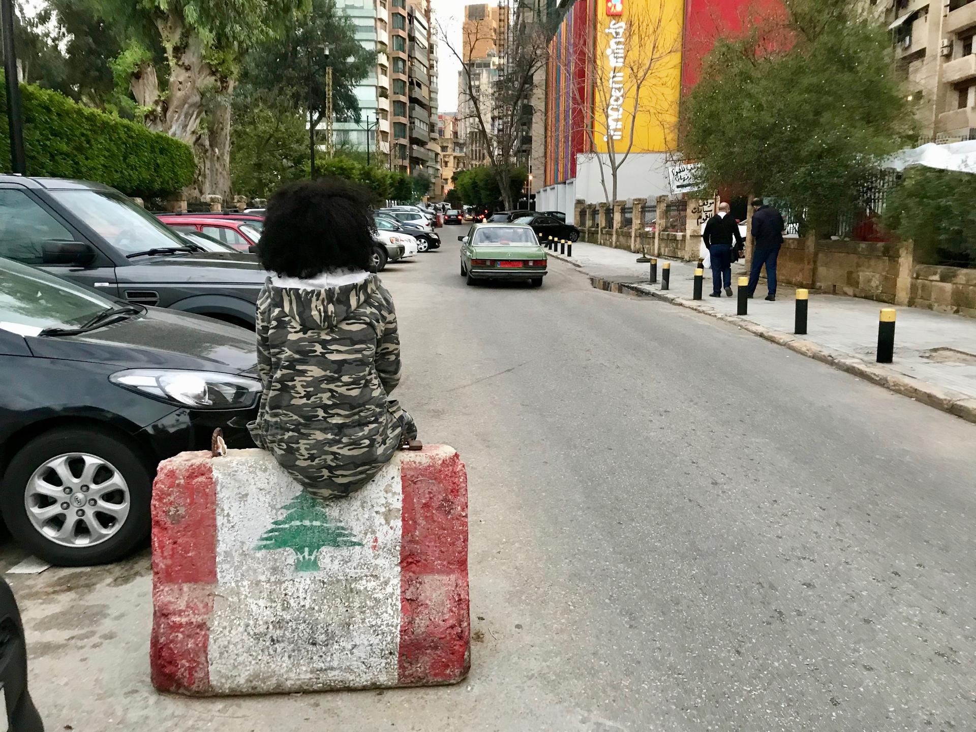 A young woman wearing a camouflage jacket sits on a barricade in middle of street.