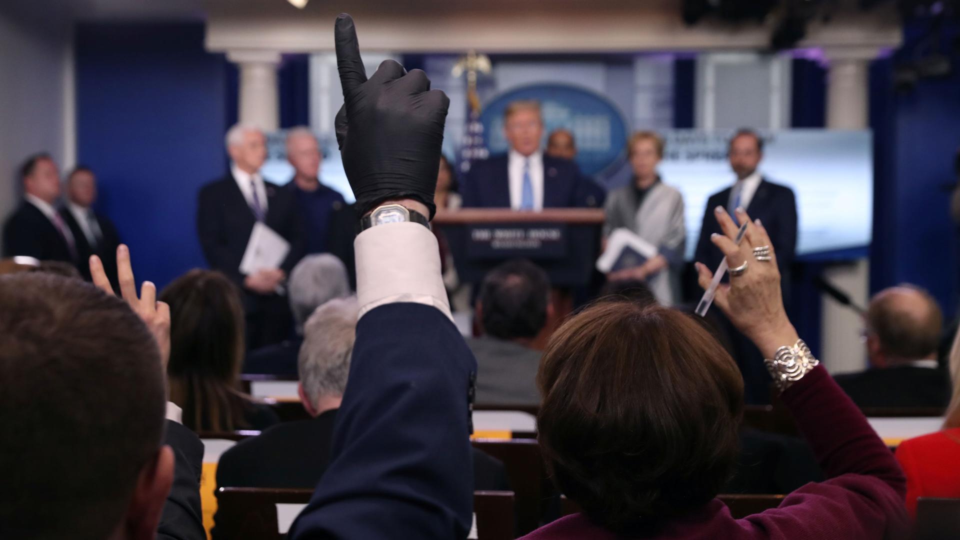 A group of reporters are shown sitting in chairs with one person holding up their hand which is covered by a dark colored latex glove.