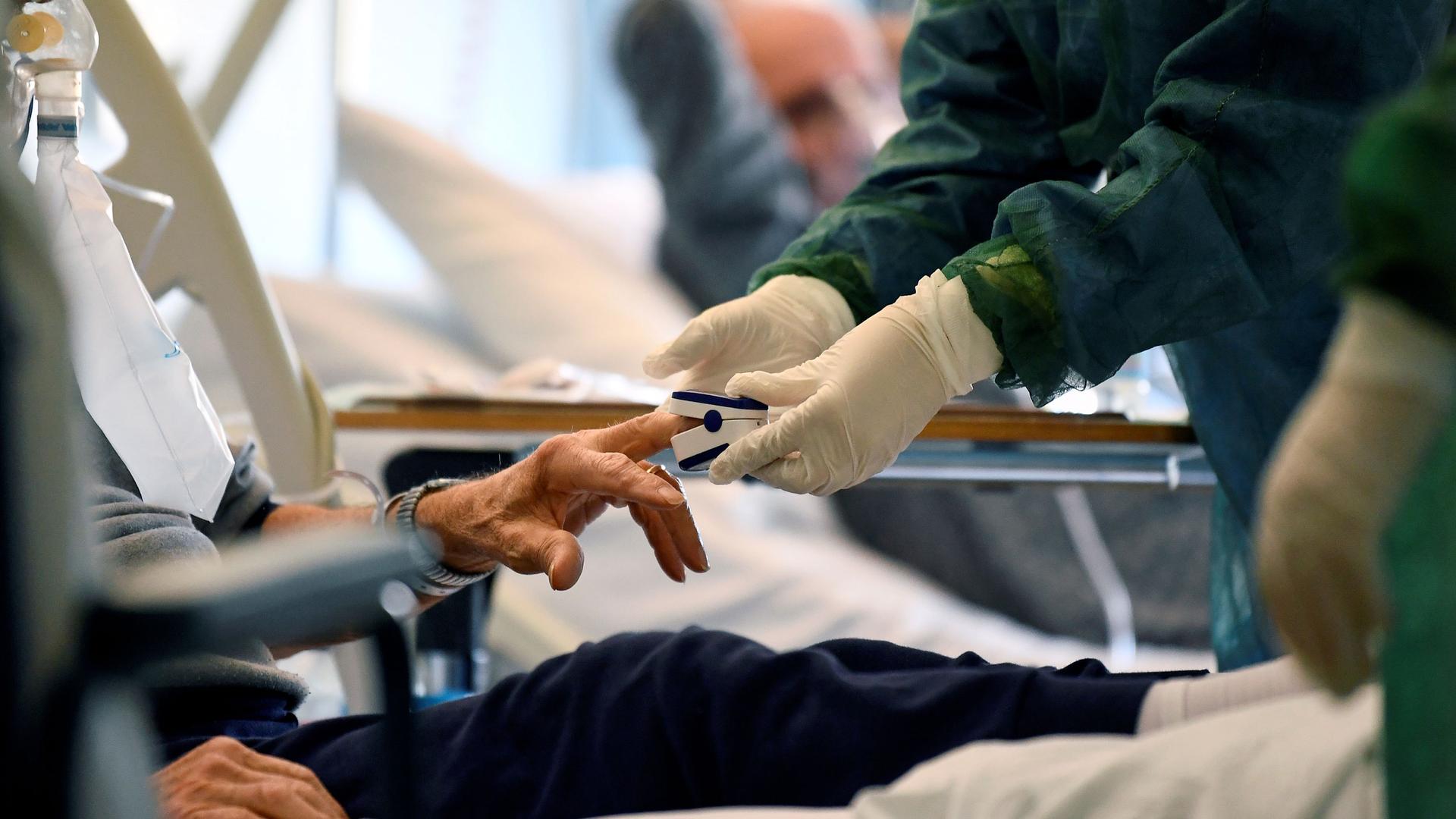 The hands of a medical professional are shown wearing protective gloves and putting a device on a patient's finger.
