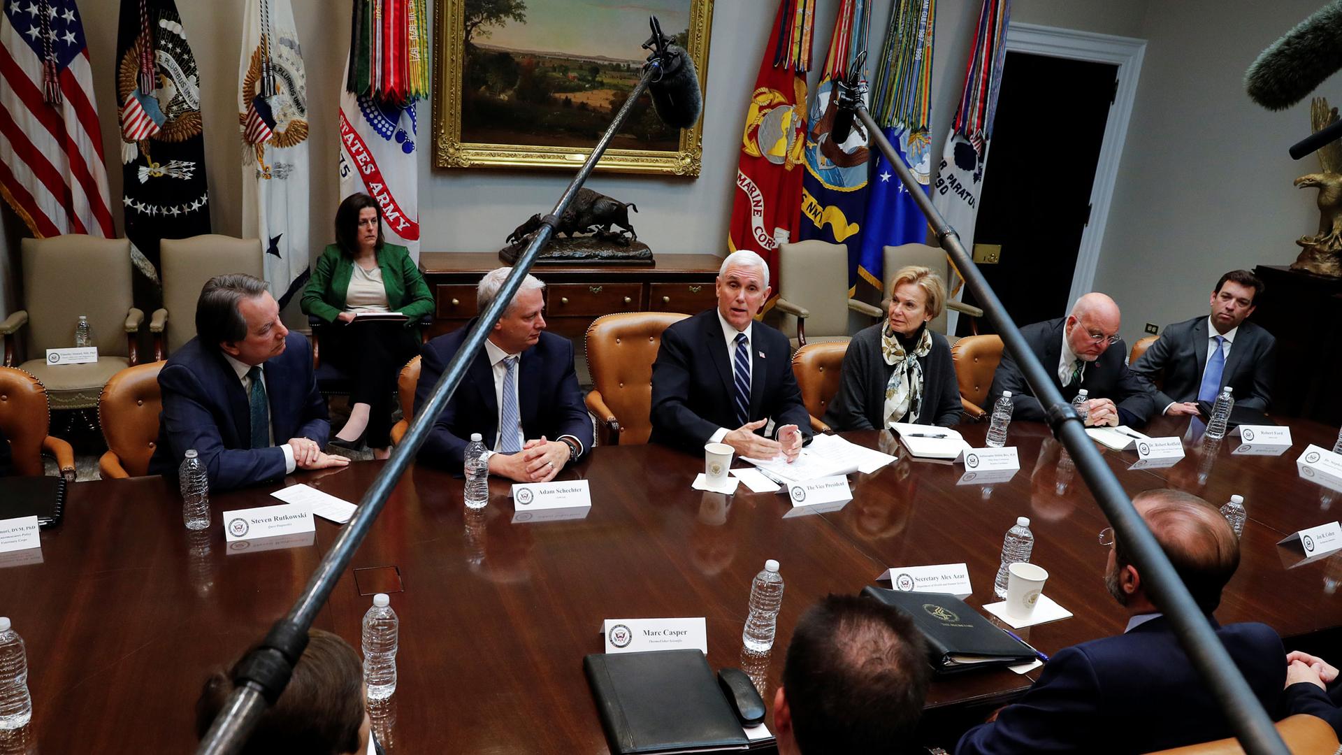 US Vice President Mike Pence is shown sitting at a table with people sitting all around him and microphones on boom polls hanging overhead.