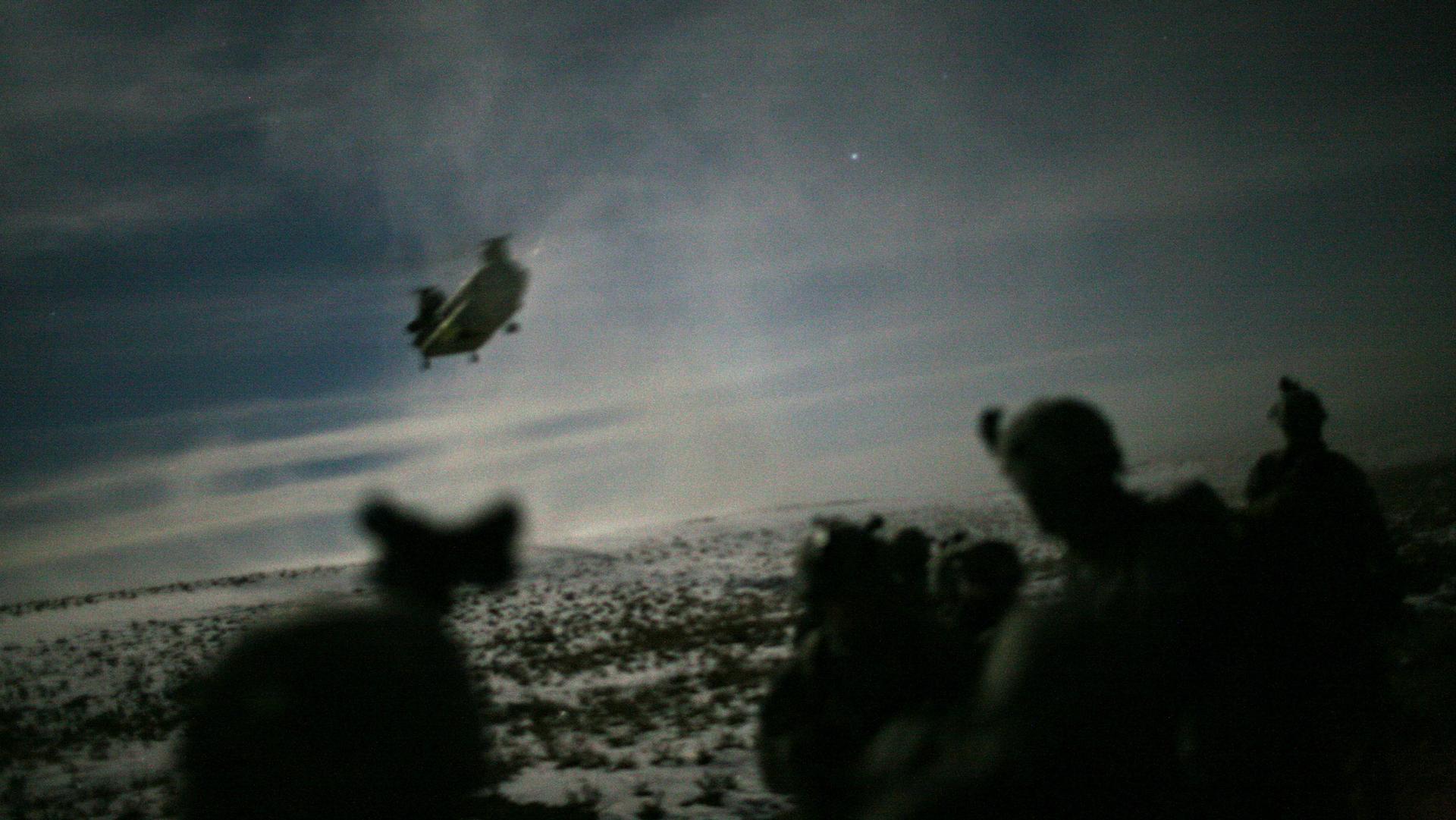 A large helicopter is shown in the distance making its landing approach with the shadow of soldiers in the nearground.
