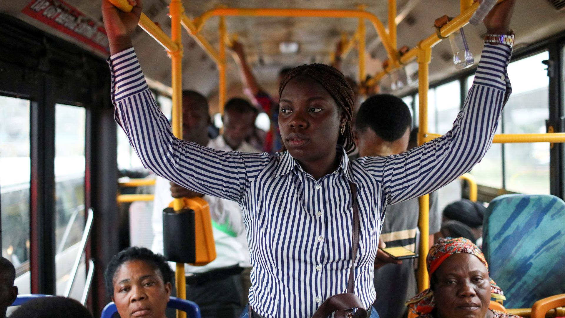 A young woman is shown standing in the middle aisle of a bus holding on to yellow handles.