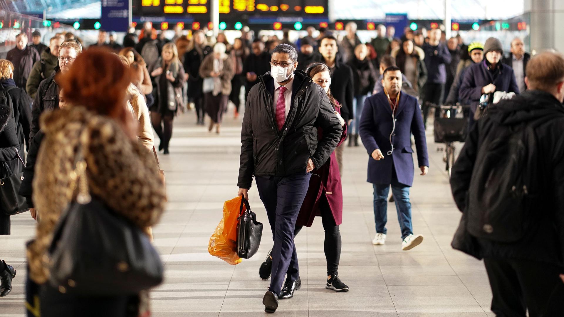 A large crowd of people are seen at Waterloo station with a man in the center of the frame wearing a face mask.