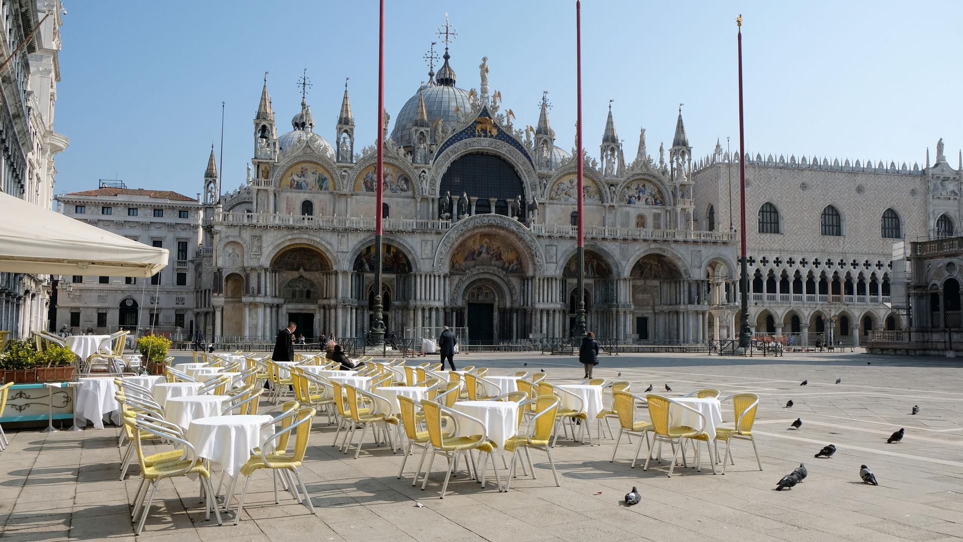 Dozen's of empty tables with white table clothes and yellow chairs are shown in St. Mark's square.