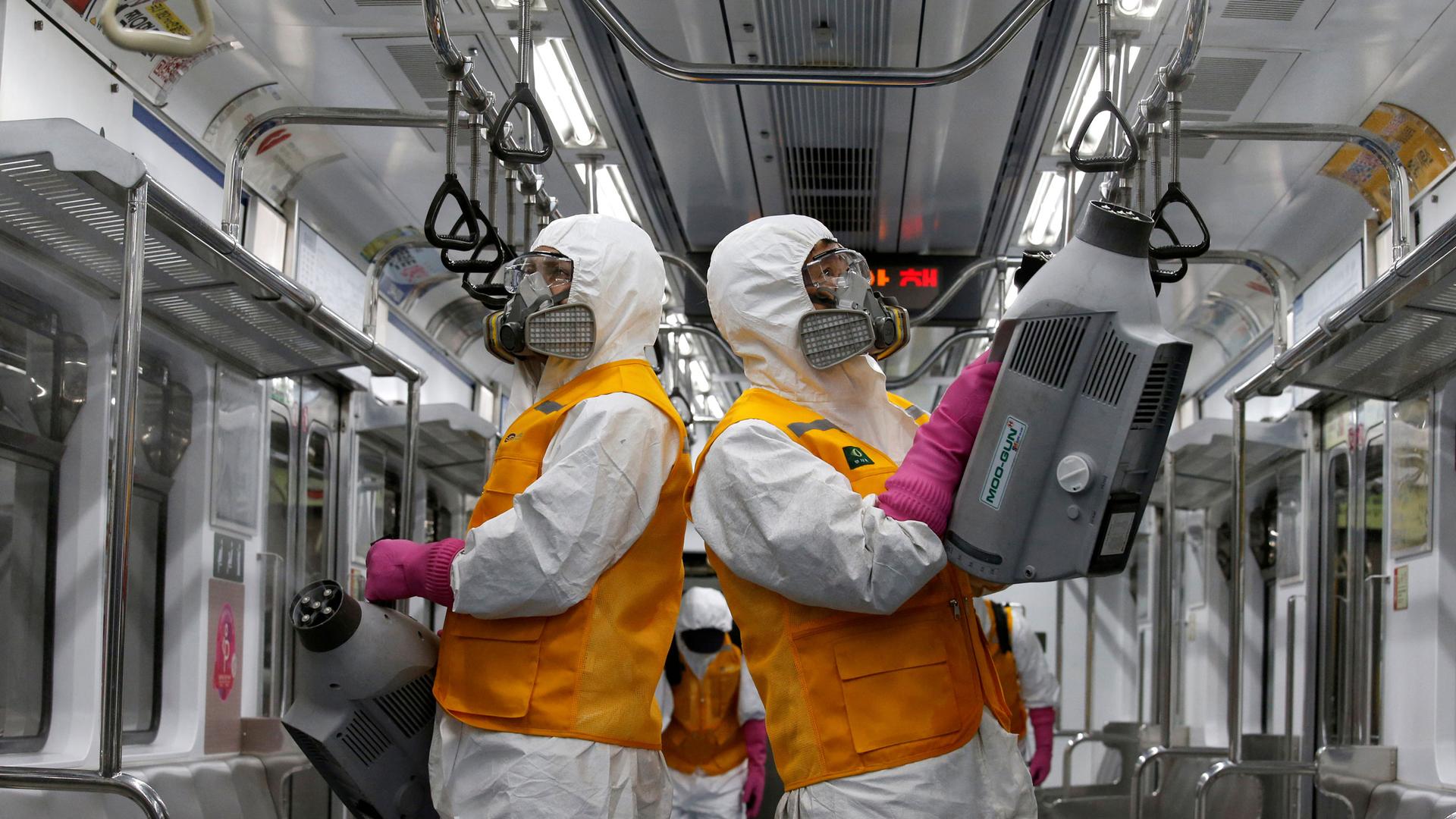 Two workers are shown inside a subway car with white and orange protective and carrying machines that are cleaning the car.