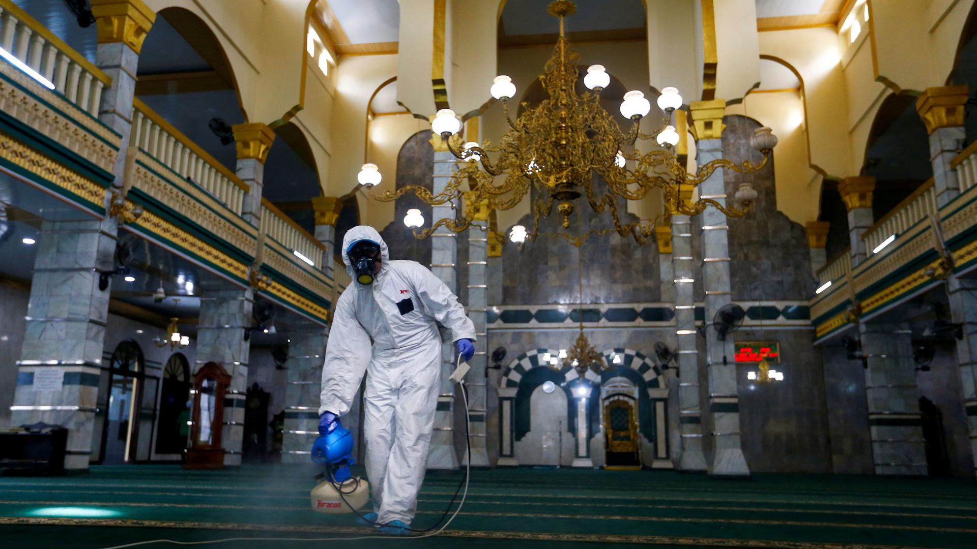 A medical officer wearing a white protective suit and a full face mask is shown spraying a carpet of a open area.