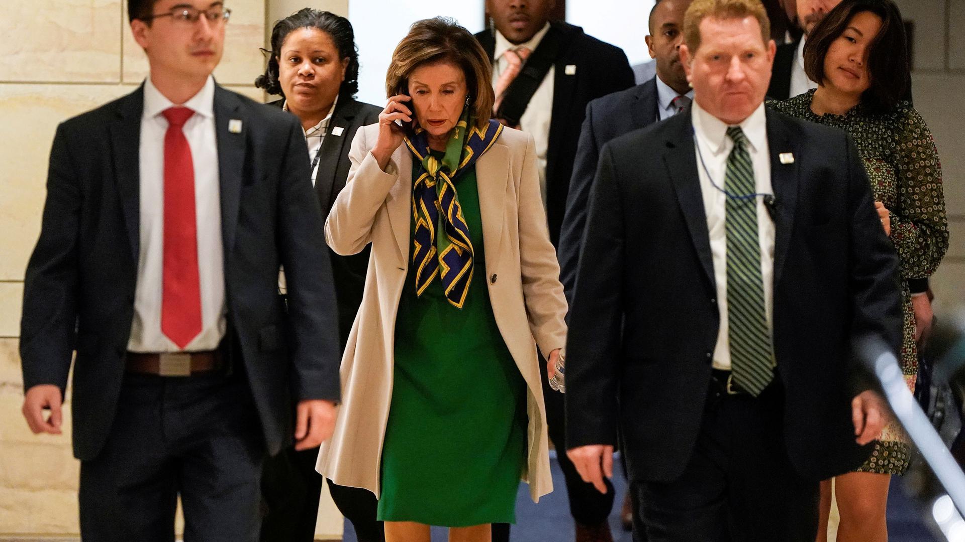 Speaker of the House Nancy Pelosi is shown walking amongst a group of people and wearing a green dress and khaki jacket while speaking on the phone.