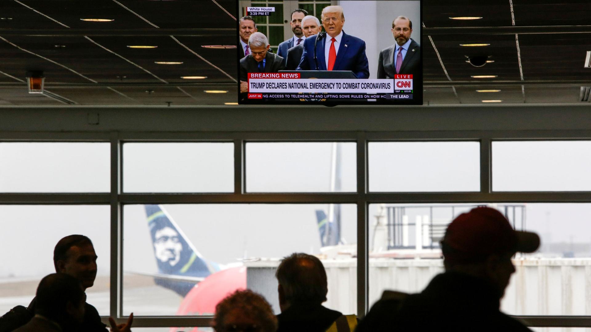 A television is shown hanging from the ceiling with US President Donald Trump displayed while several people are in shadow below.
