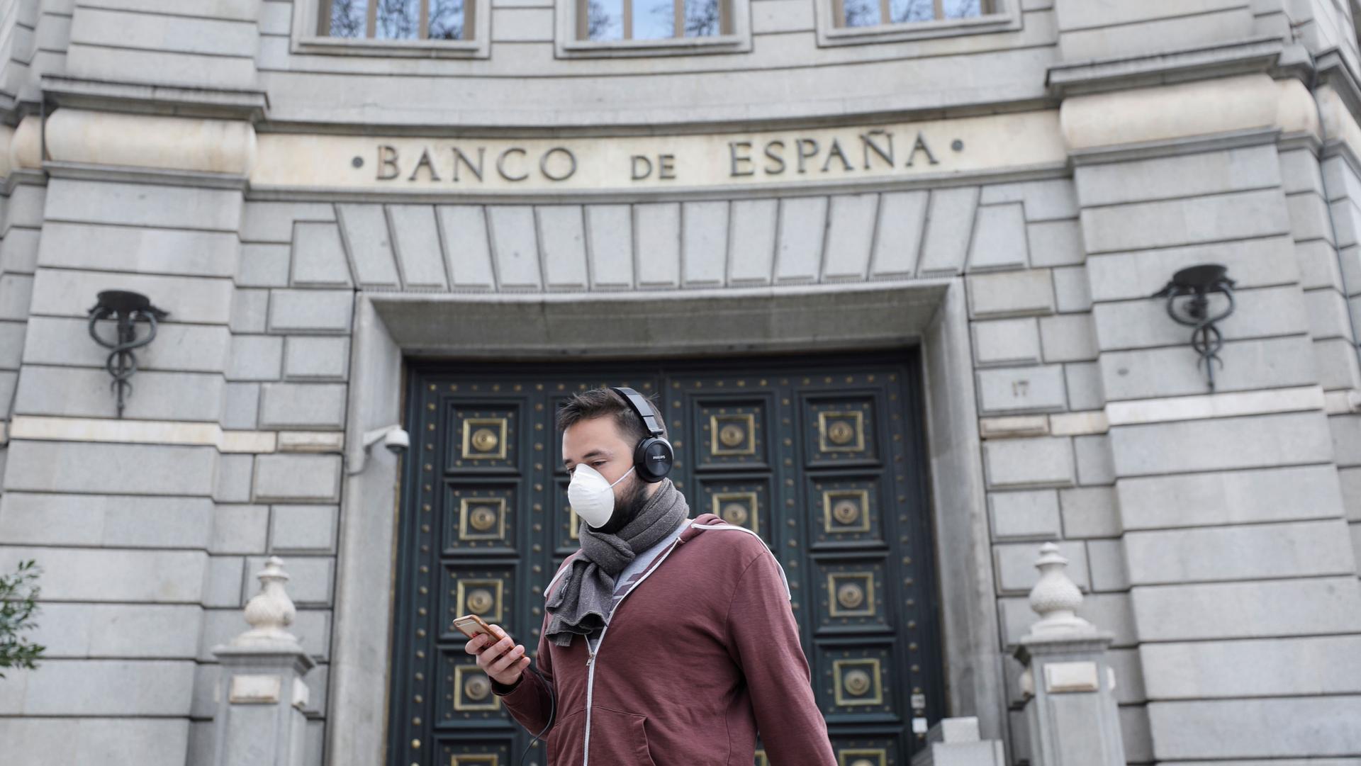 A man is shown wearing a medical face mask and large headphones while looking at a mobile phone.