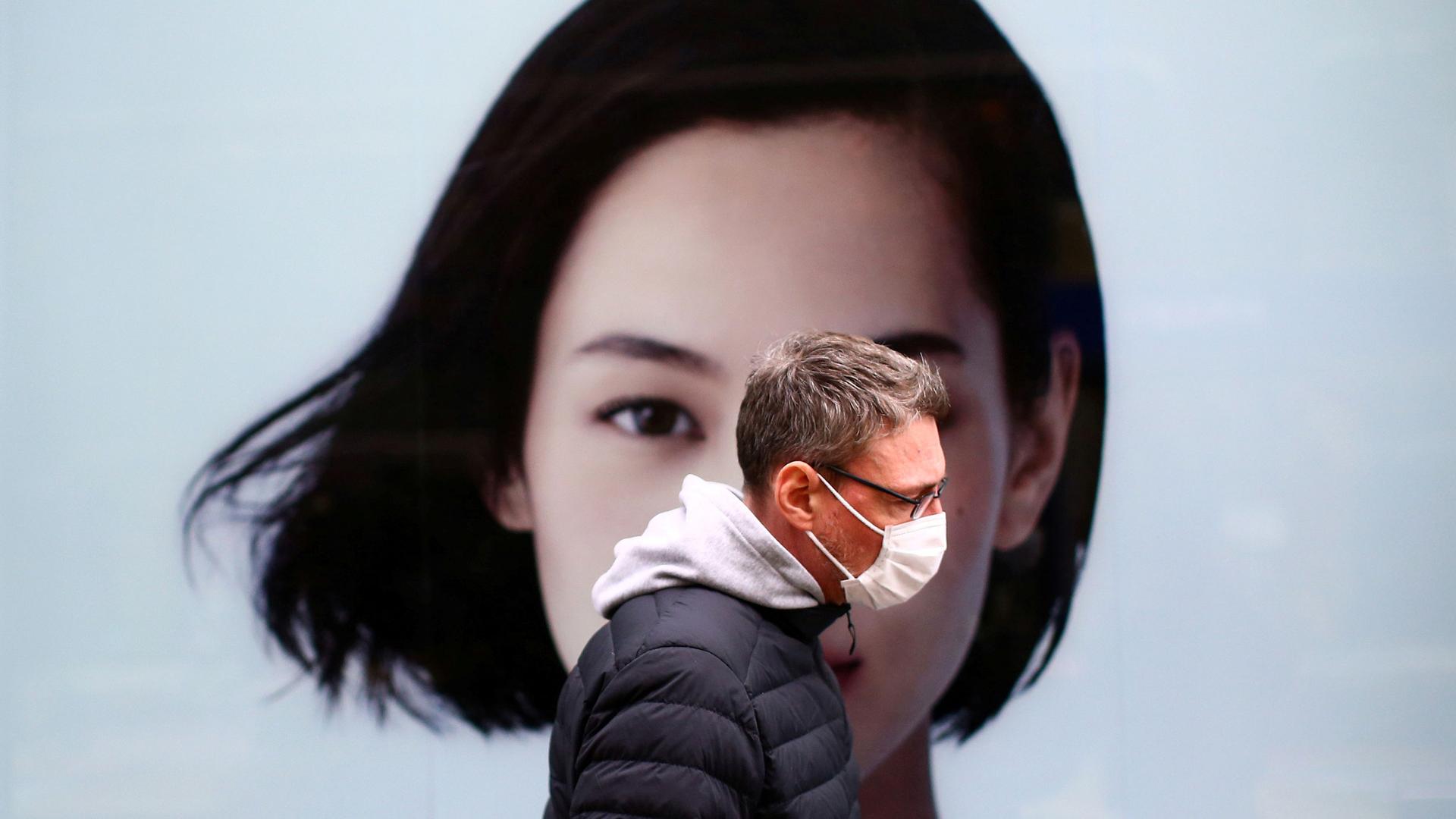 A man is shown wearing a puff jacket and face mask and walking past a advertisement with the face of a woman on it.