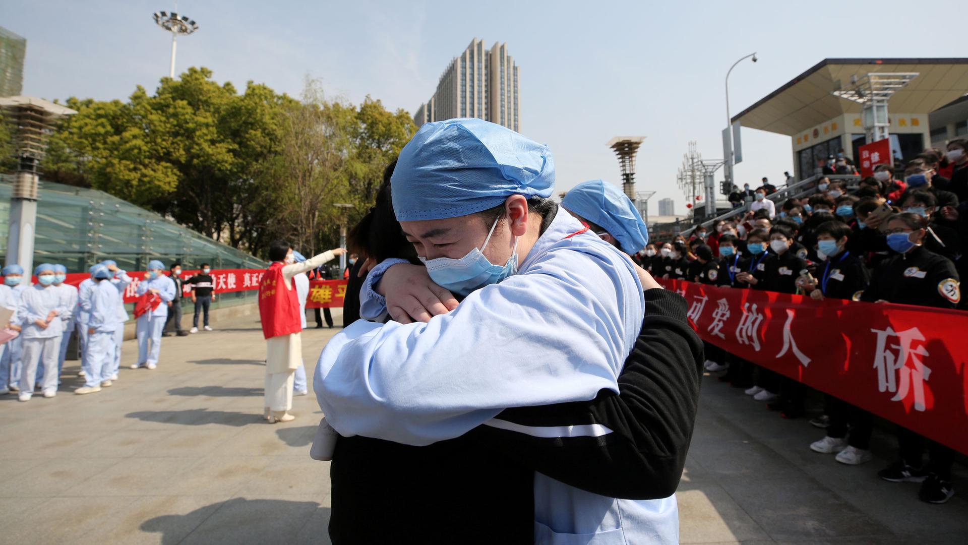 A man wearing a face mask and protective clothing is shown hugging another person in the street.