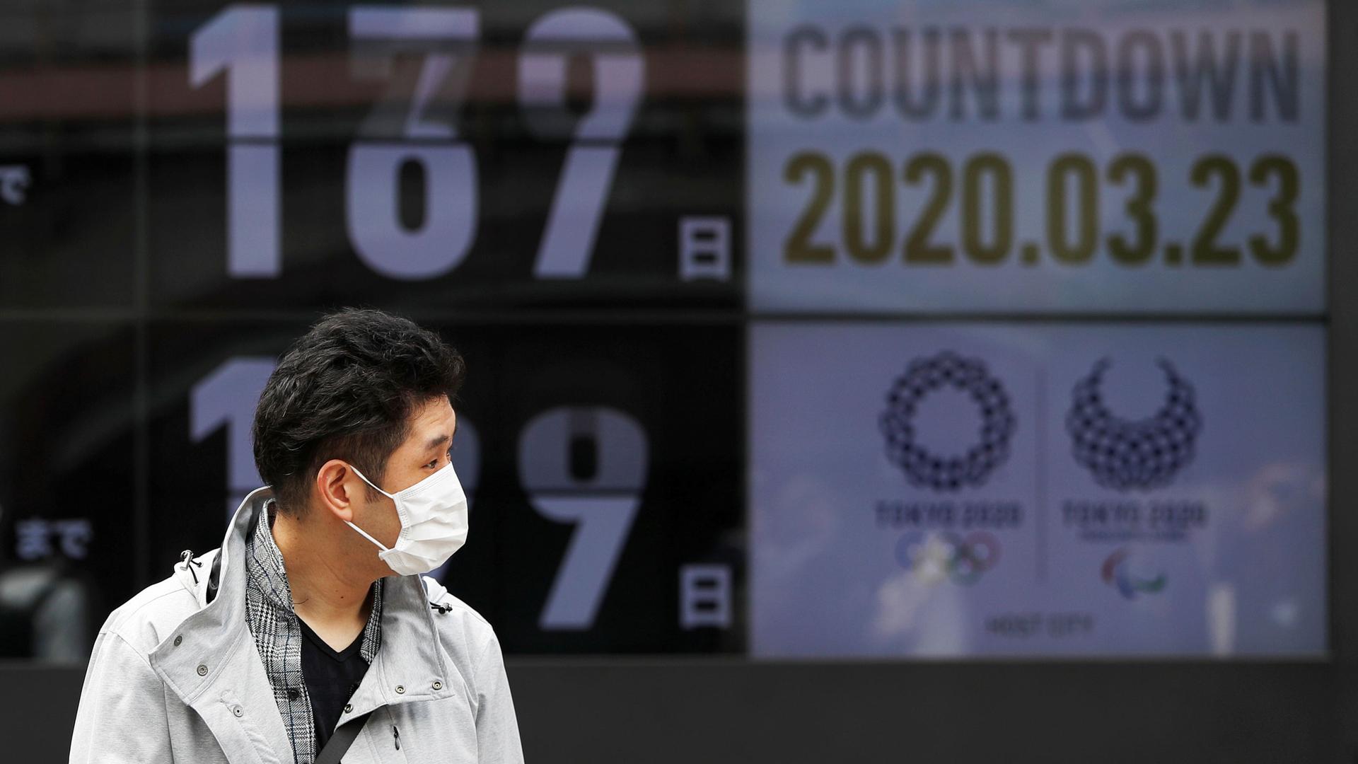 A man is shown wearing a face mask and walking in front of a countdown clock for the 2020 Olympics Games in Tokyo.