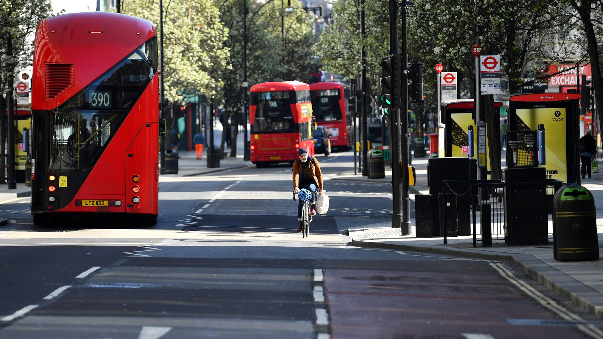 Three red, double-decker buses are shown driving on a nearly empty street that also includes a lone bicyclist.