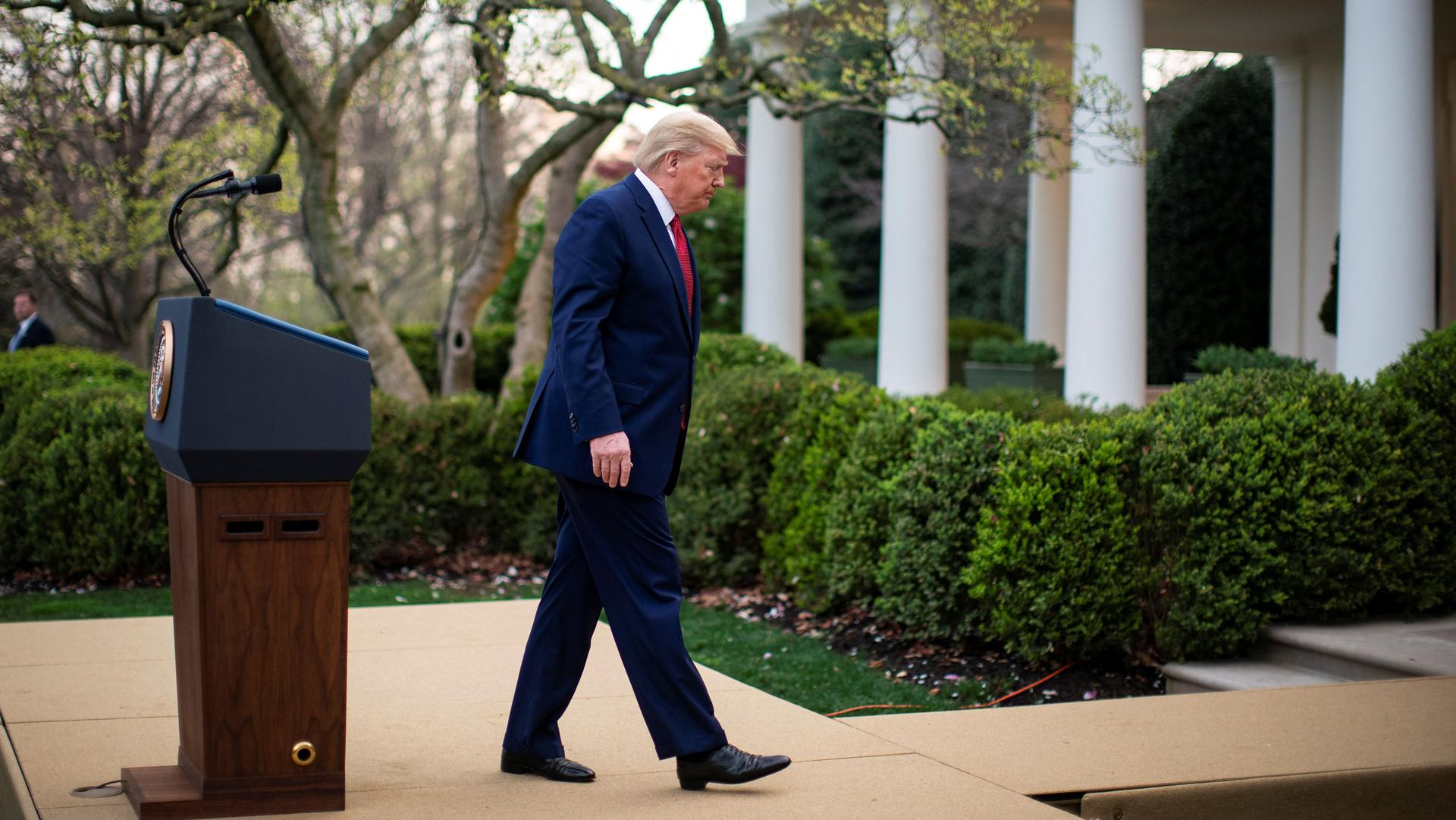 US President Donald Trump is shown wearing a blue suit and walking away from a wooden podium at the White House.