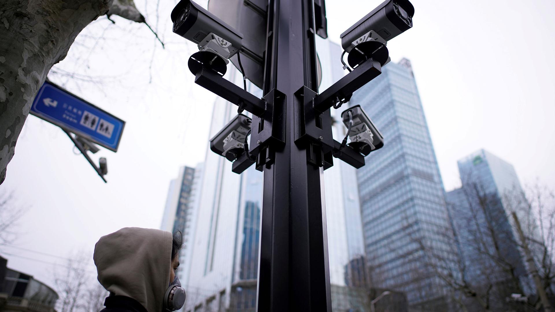 A metal poll is shown with four surveillance cameras pointed out in four separate directions as a man walks by wearing a face mask.