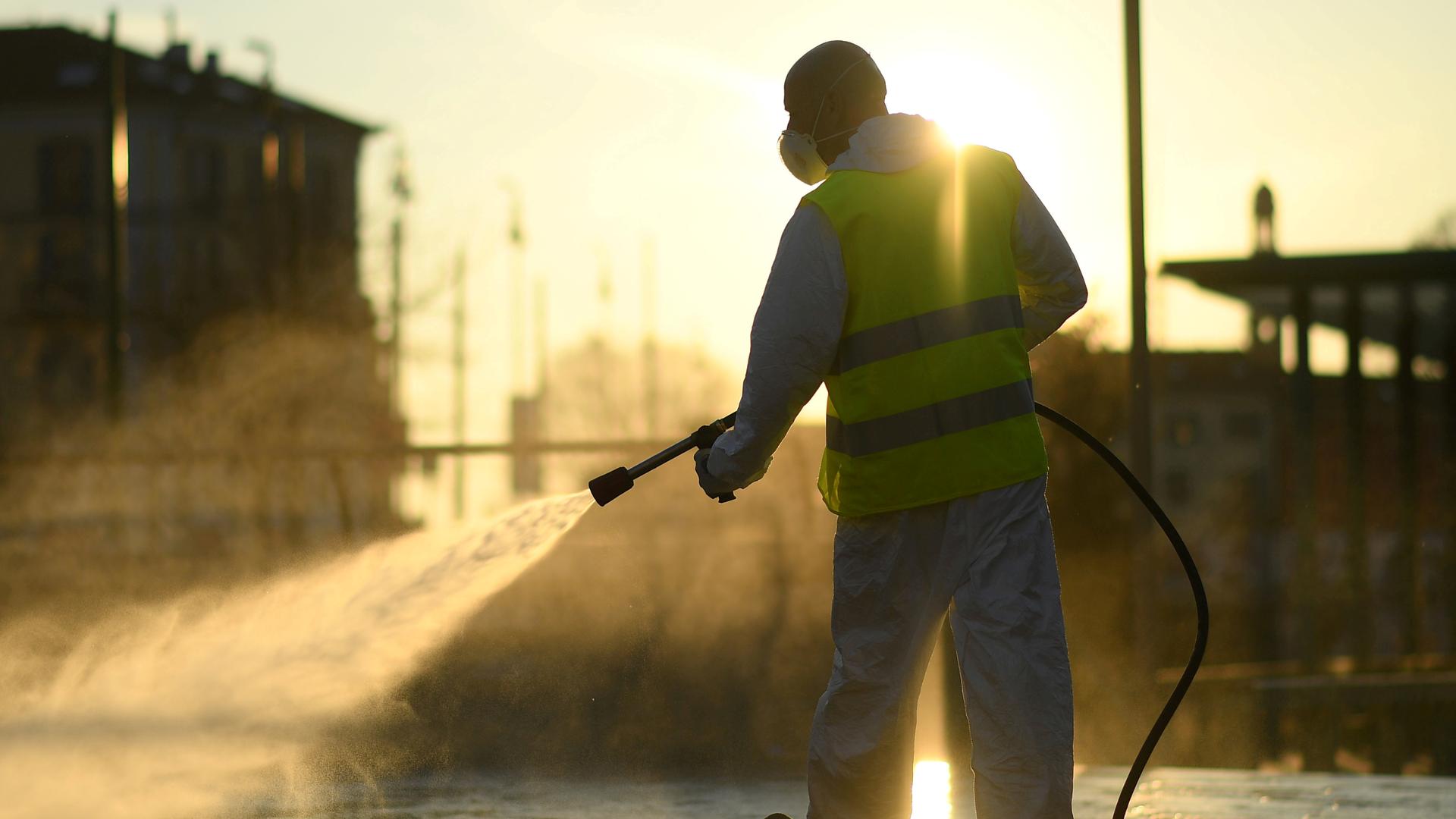 A man sprays a street with sun streaming in as he wears protective gear. 