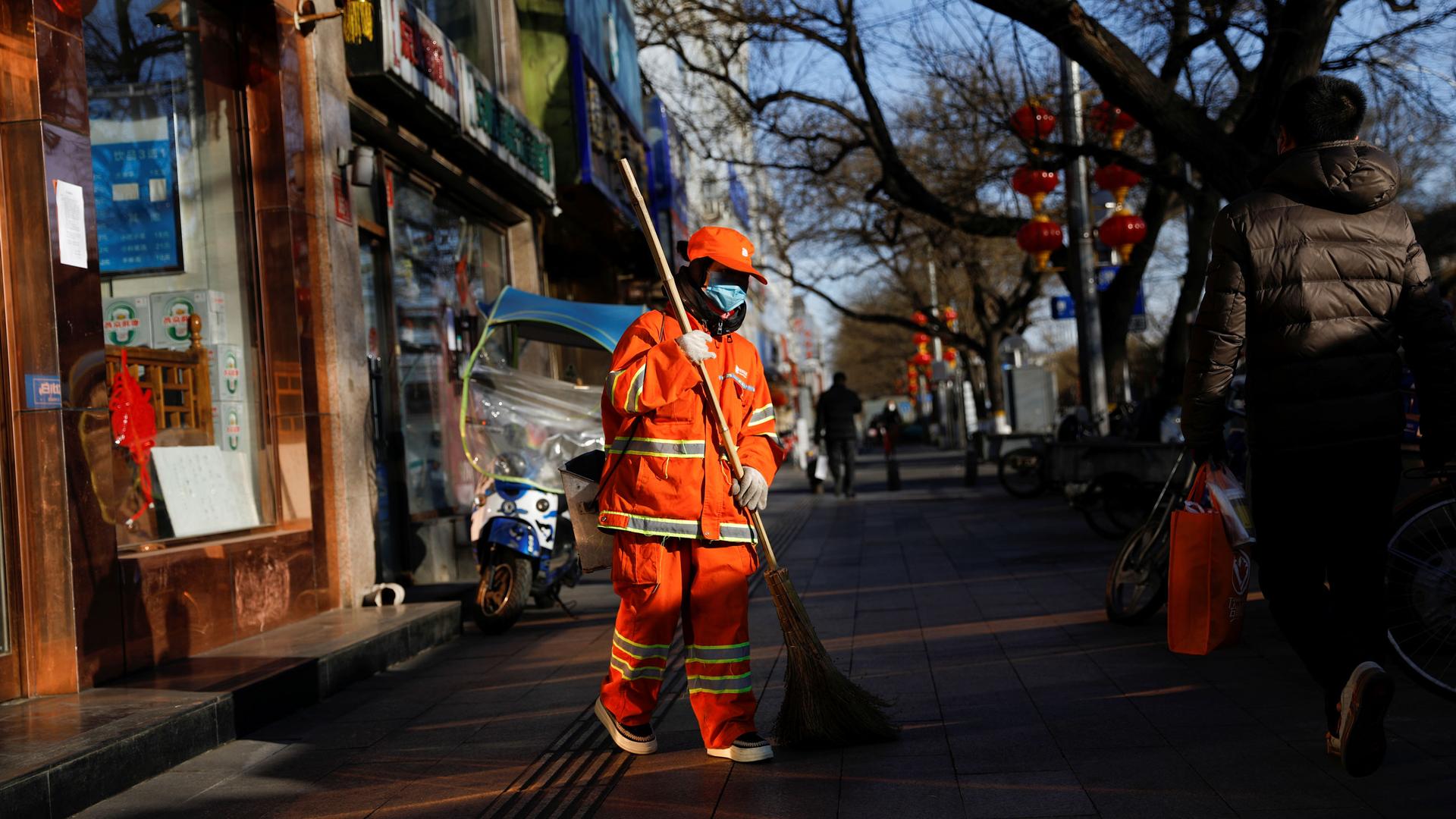 A street cleaner is shown wearing an orange, reflective safety suit and a face mask walks on a sidewalk.