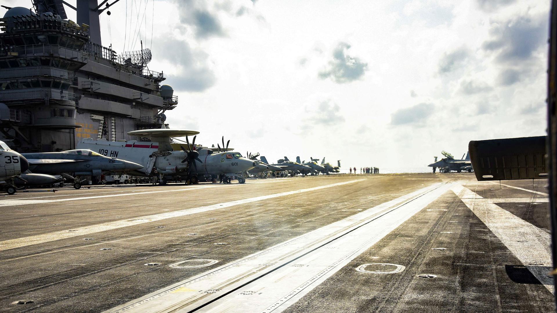 A photograph taken from the deck of the USS Theodore Roosevelt aircraft carrier with the ship's tower and several airplanes in the background.