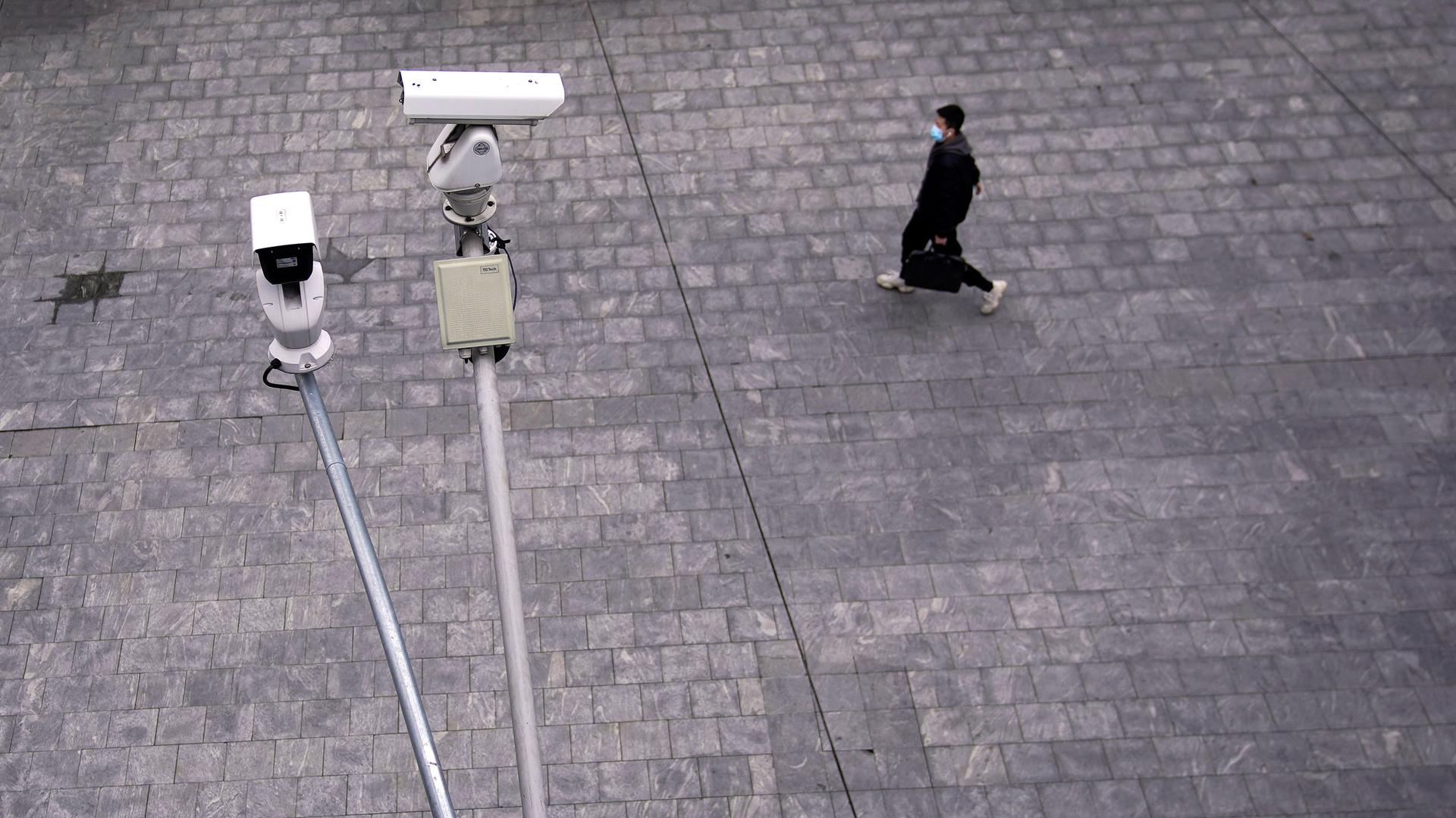 Two surveillance cameras are shown in the nearground with a man walking in a stone walkway and wearing a protective face mask in the distance.