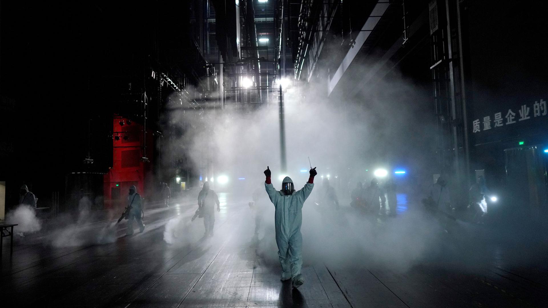 Several people are shown wearing protective clothing and spraying disinfectant in an open area in the dark of the evening.