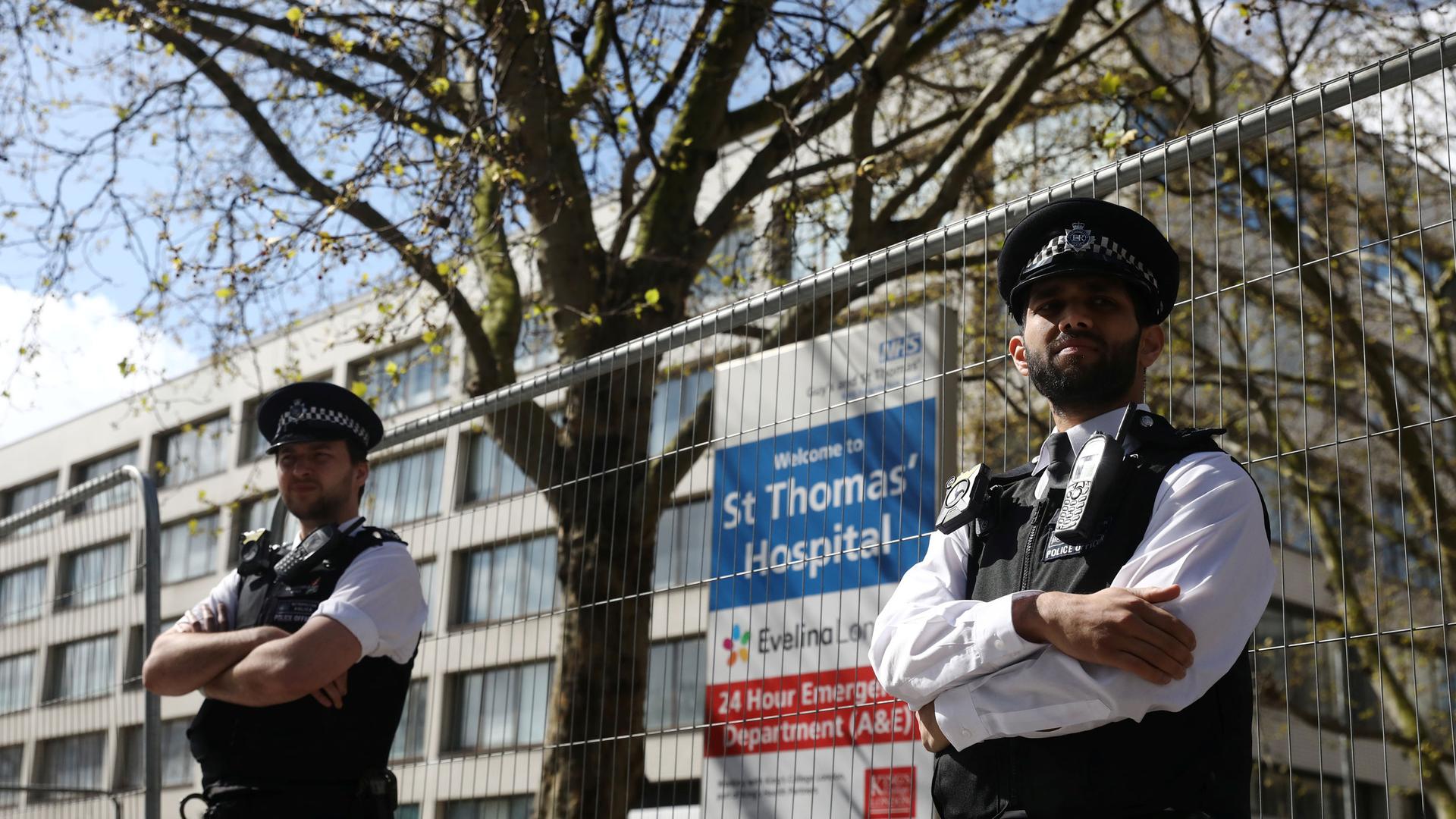 Two police officers are shown with their arms folded and standing in front of a fence with St Thomas' Hospital  in the background.