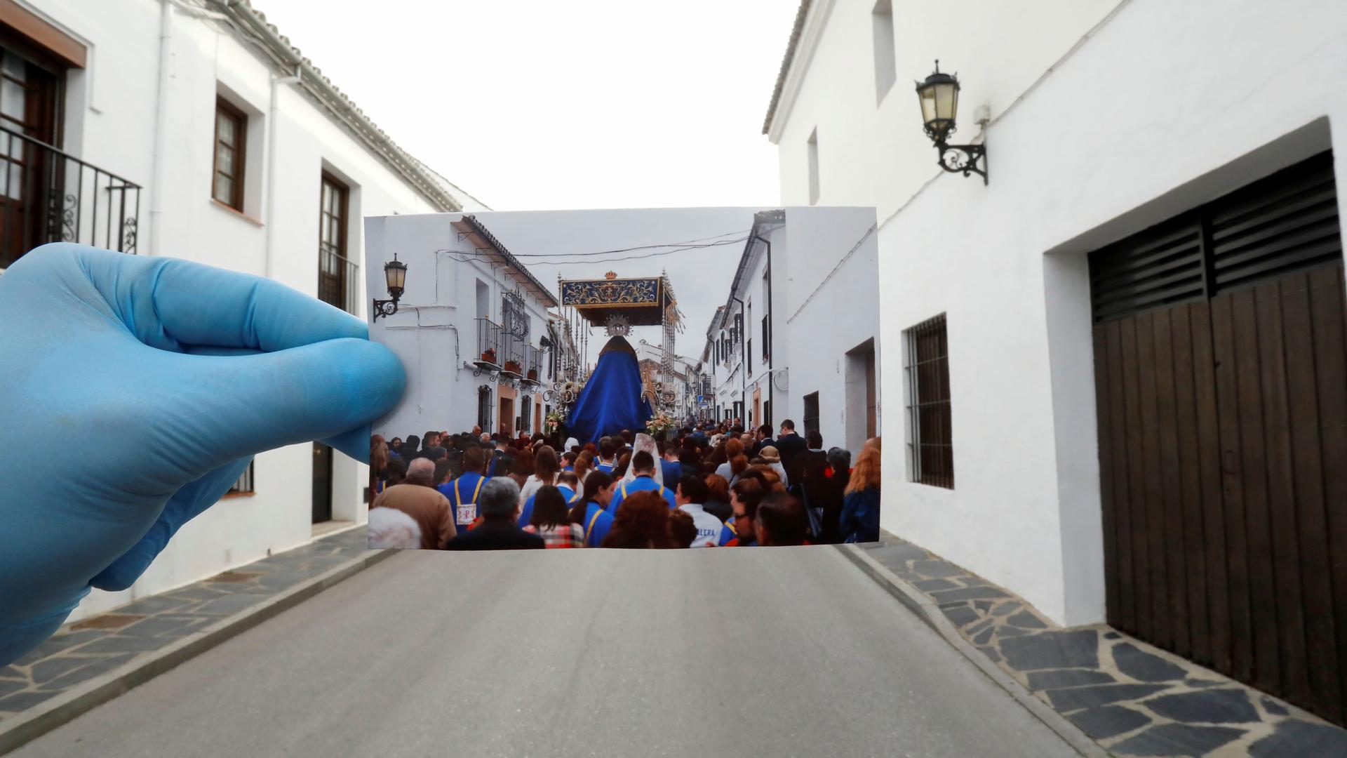 A photographer wearing a protective blue glove and is holding a picture of the very street they are standing in — presently empty and in the photo filled with people.